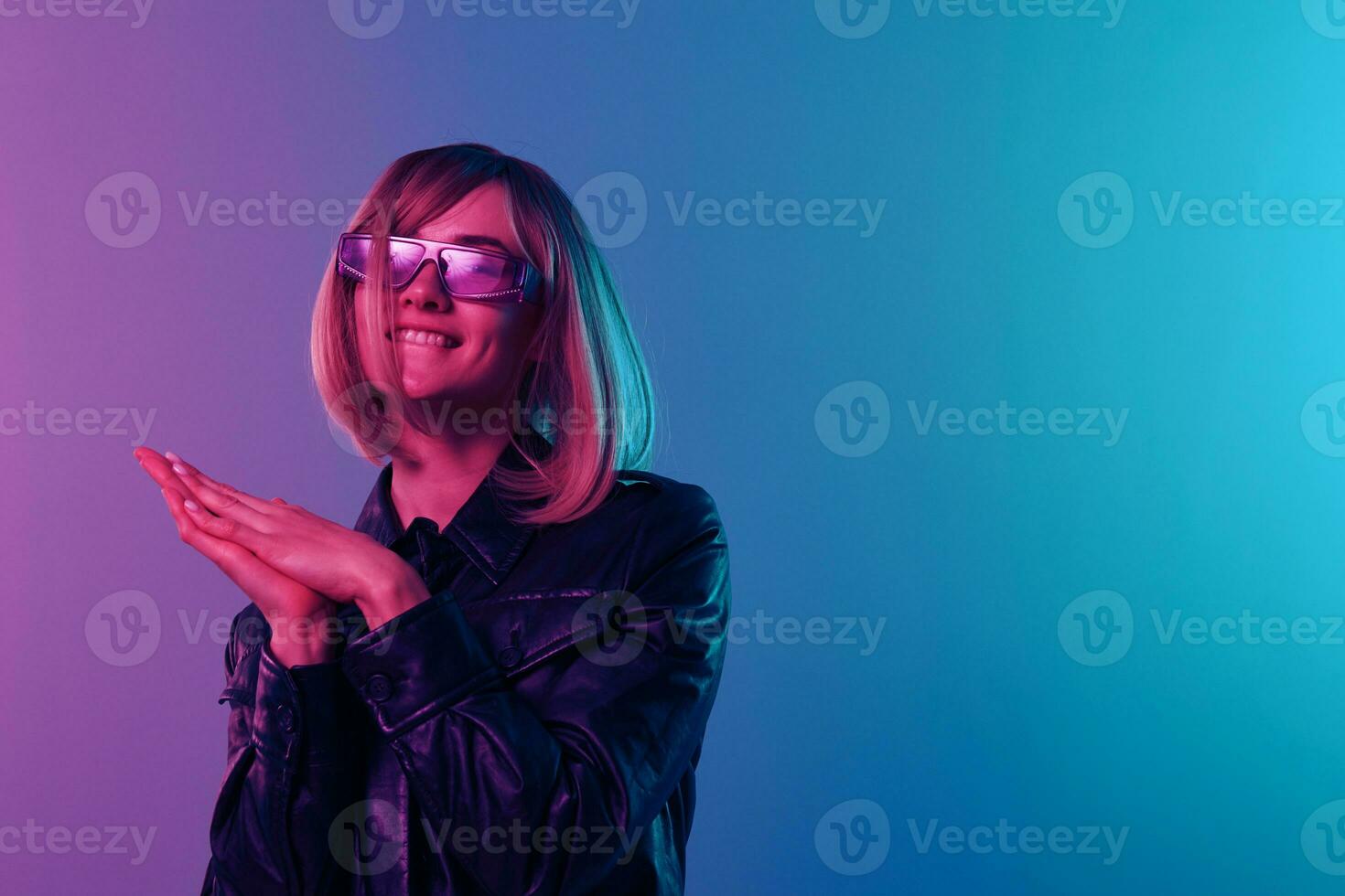 Cute beautiful blonde woman in leather jacket sparkly dress trendy sunglasses fold hands look at camera posing isolated in blue pink color light studio background. Neon party concept. Copy space photo