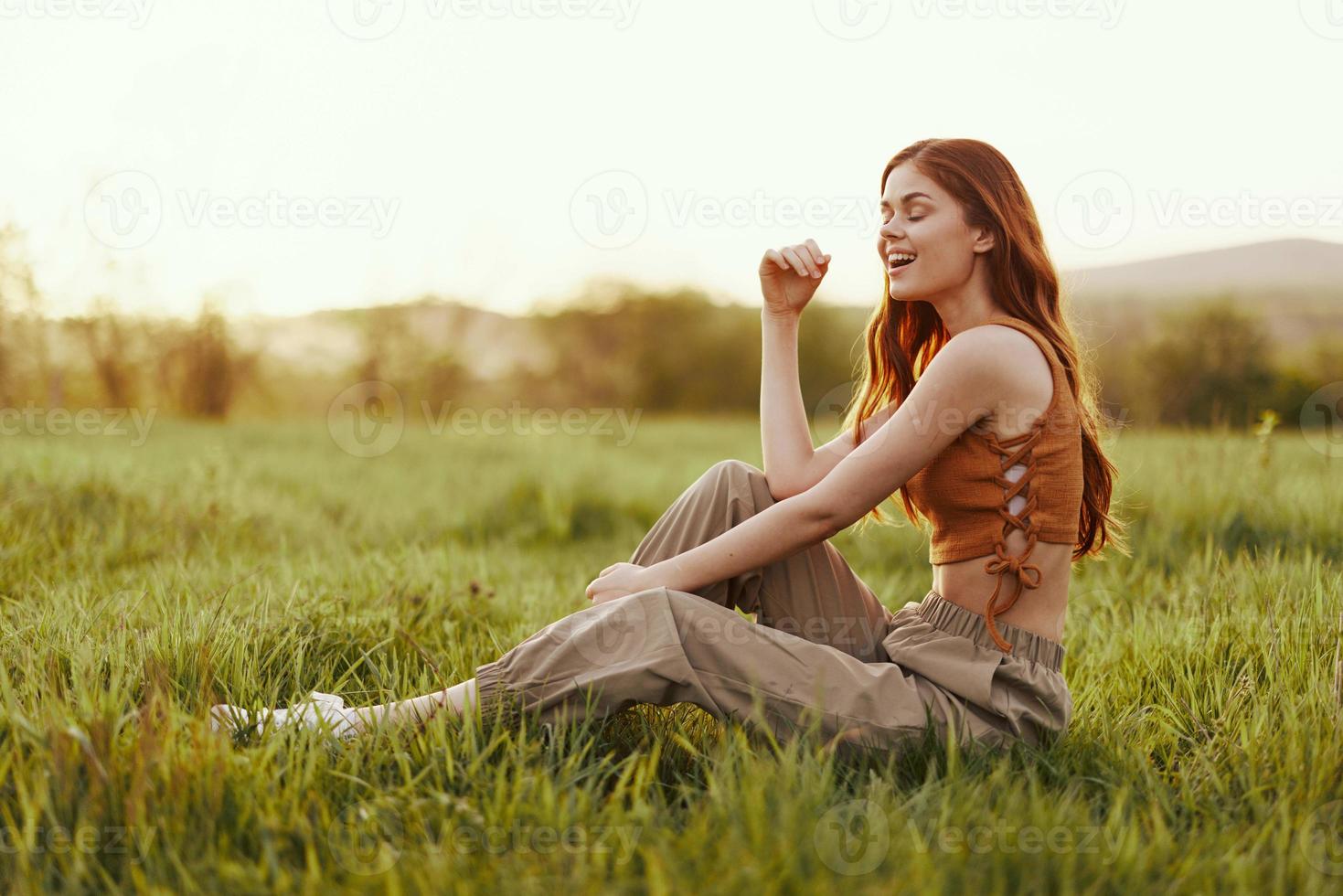 un mujer se sienta en el verde joven Fresco césped en el abierto aire y se ríe y sonrisas felizmente en el puesta de sol luz, su volador rojo pelo revoloteando en el viento foto