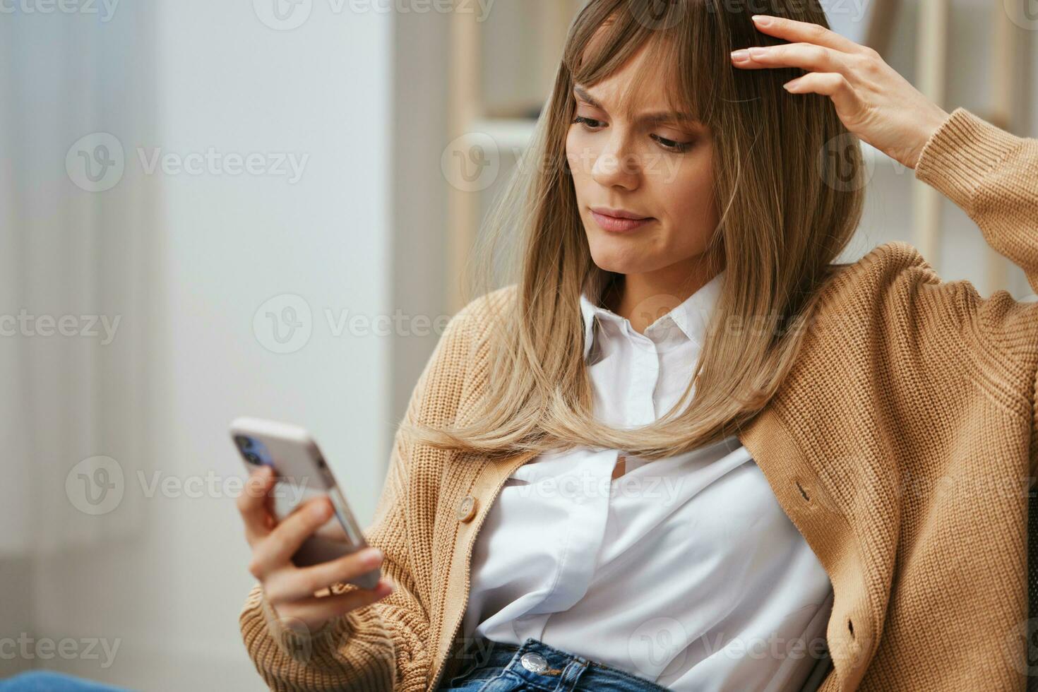 Nervous thoughtful young blonde lady in warm sweater worried about exam results use phone sitting in armchair at home. Pause from work, take a break, social media in free time concept. Copy space photo