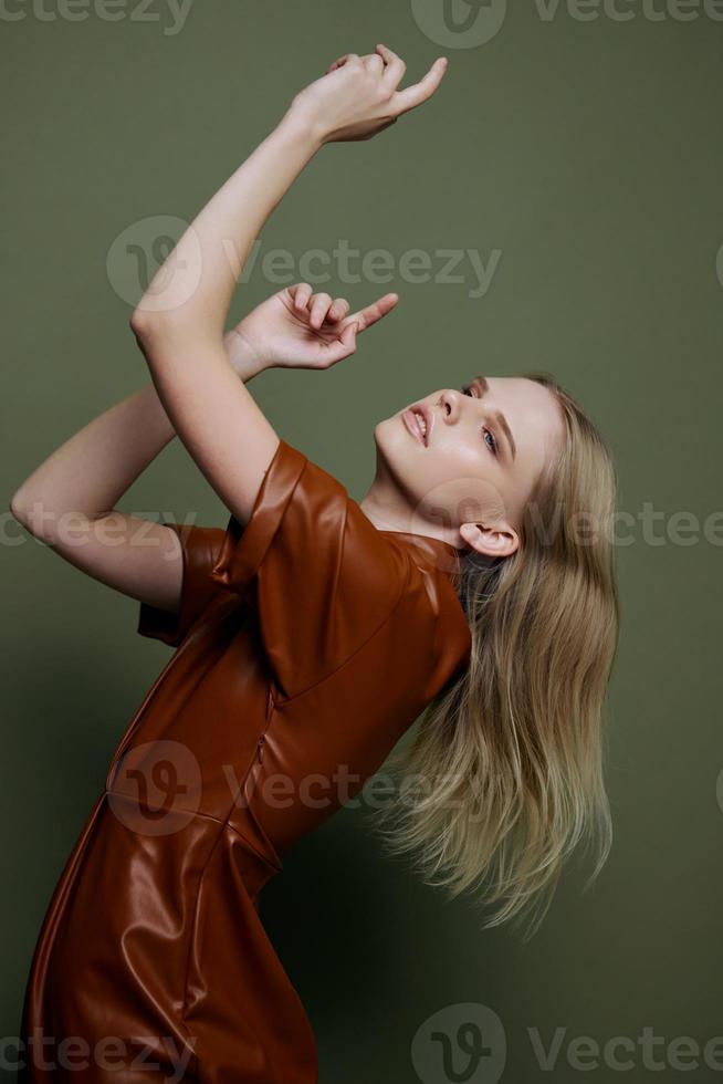 Curving in dance blonde young female model in brown trendy dress holding hands up dreaming looks up aside posing isolated on over olive green studio wall background. Seasonal sale banner photo