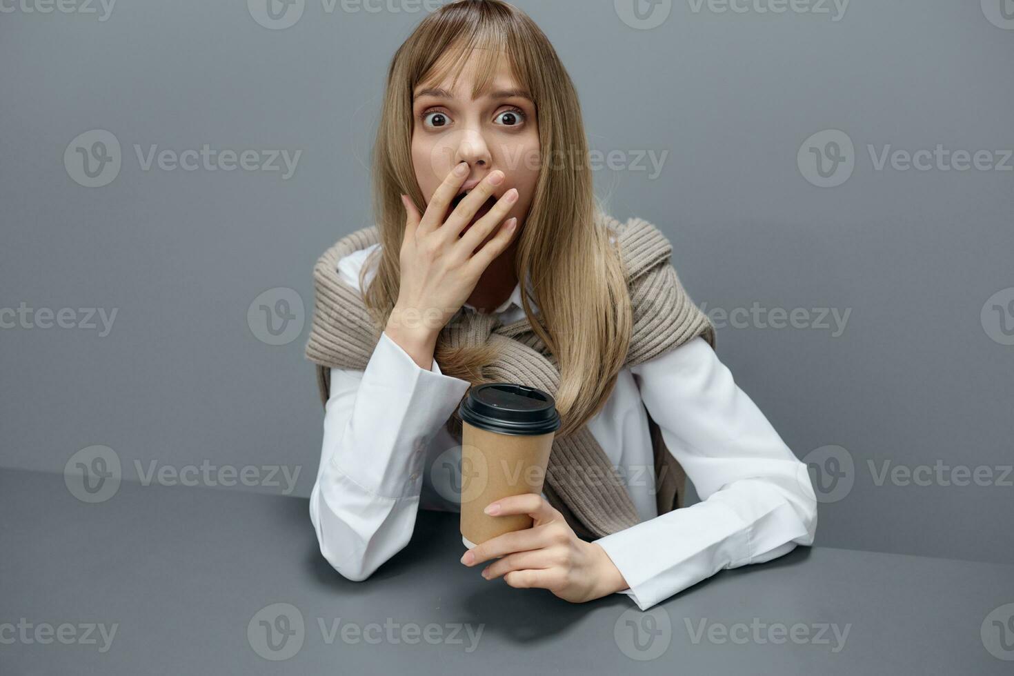 Shocked young blonde student lady freelancer in warm sweater hold takeaway coffee close mouth sitting at workplace in gray modern home office. Coffee break Lover Concept. Copy space. Cool offer photo