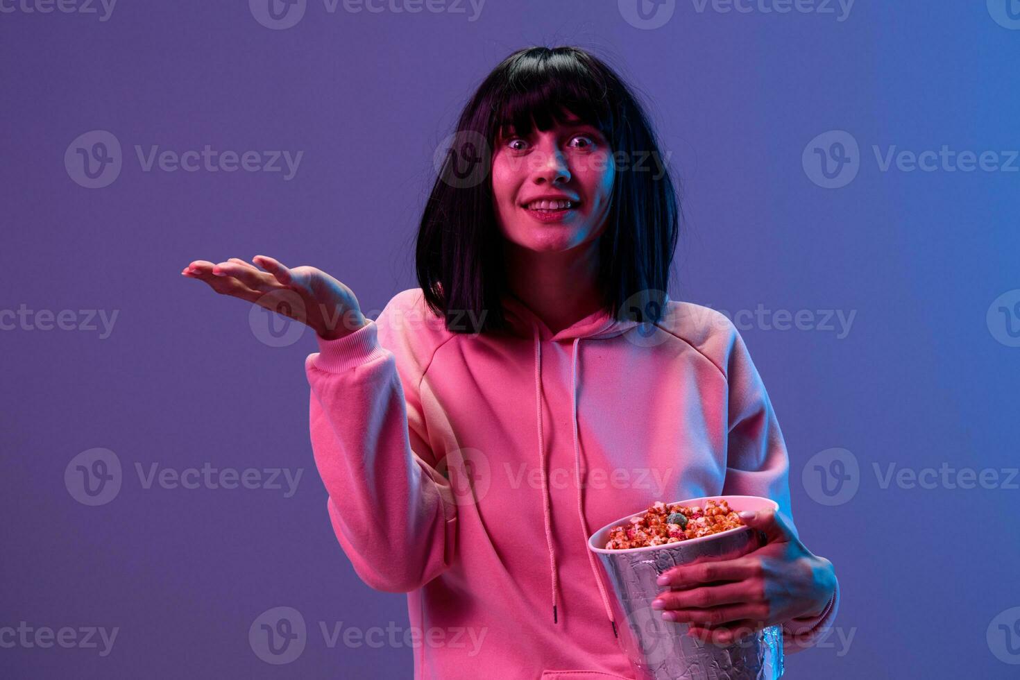 Happy shocked awesome brunet woman in pink hoodie with popcorn hold palm up posing isolated in blue violet color light studio background. Neon party Cyberpunk Cinema concept. Copy space photo