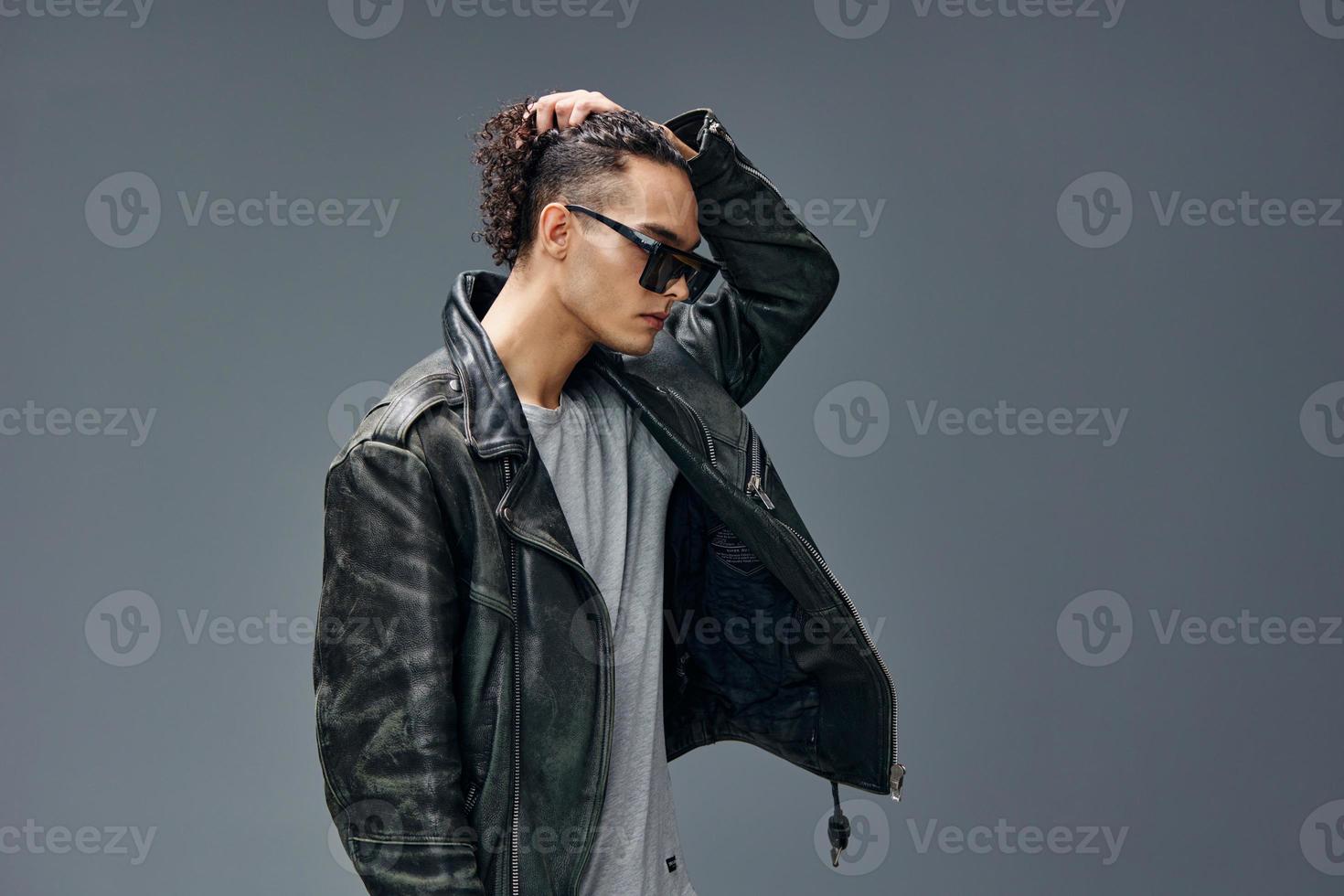 Side of view of stylish tanned curly man leather jacket trendy sunglasses posing isolated on gray studio background. Cool fashion offer. Huge Seasonal Sale New Collection concept. Copy space for ad photo