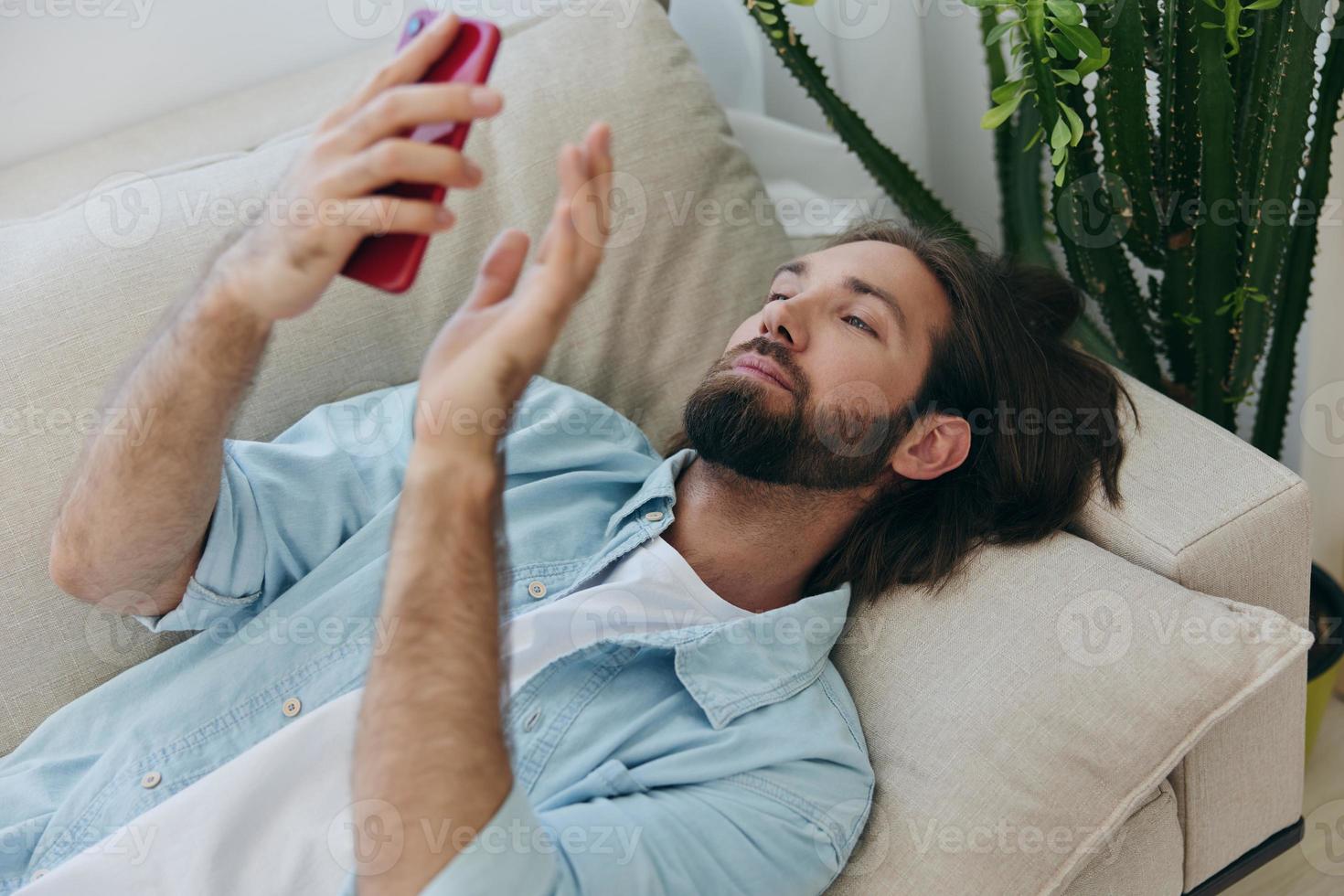 un hombre con un barba mentiras en el sofá durante el día a hogar y mira a su teléfono relajante en su día apagado, un hombre juego en el valores mercado en línea en su teléfono foto