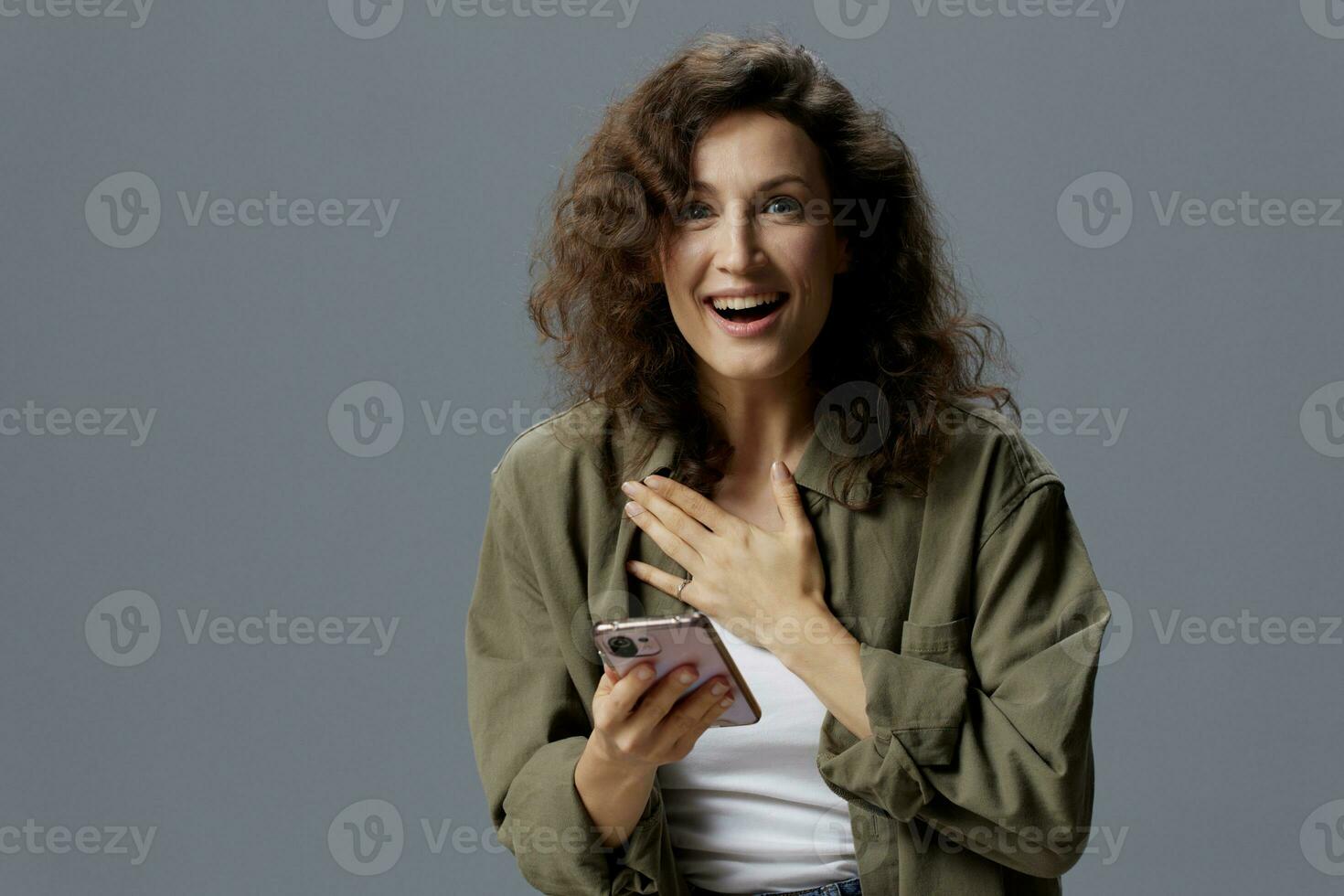 Shocked cheerful excited curly woman in casual khaki shirt received message about major sale of NFT project using phone posing isolated on gray background. Social Media Influencer concept. Copy space photo