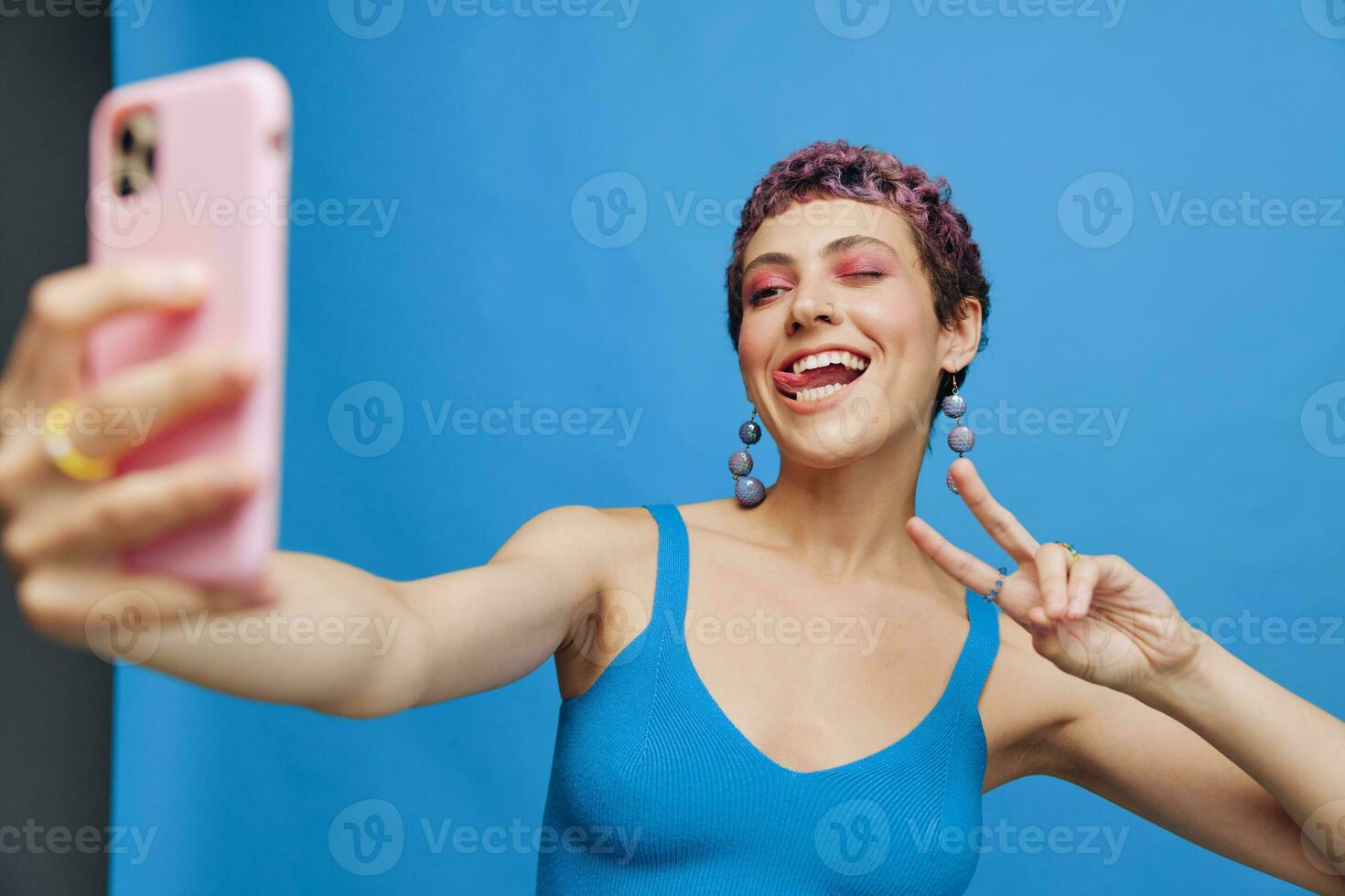 Young sports woman fashion blogger takes a picture of herself on the phone in blue sportswear smiling and showing her tongue on a blue monochrome background photo