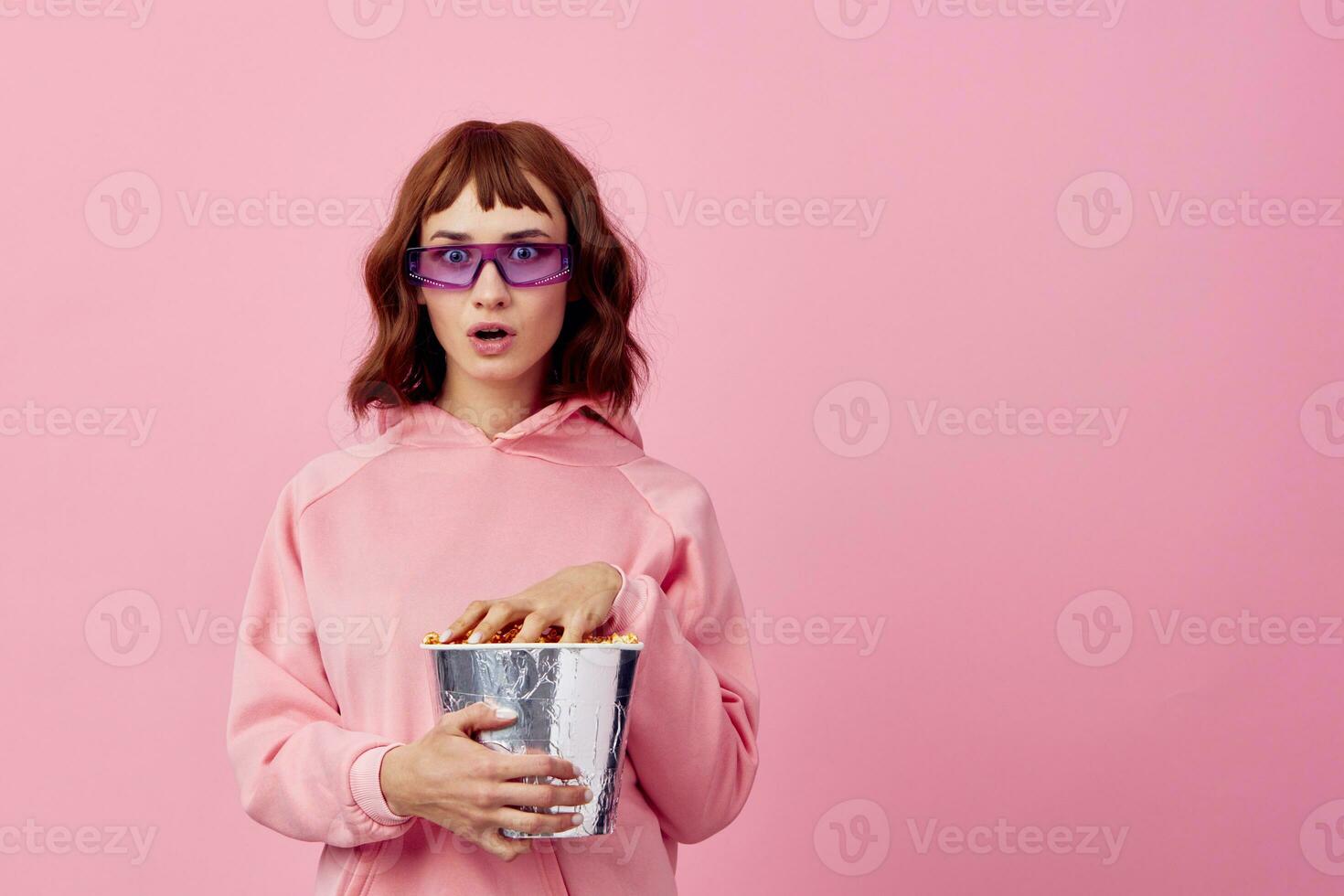 frio película. confuso linda pelirrojo dama en rosado capucha Gafas de sol con palomitas de maiz posando aislado en terminado rosado estudio antecedentes espectáculo Copiar espacio bandera. Moda cine concepto. entretenimiento oferta foto