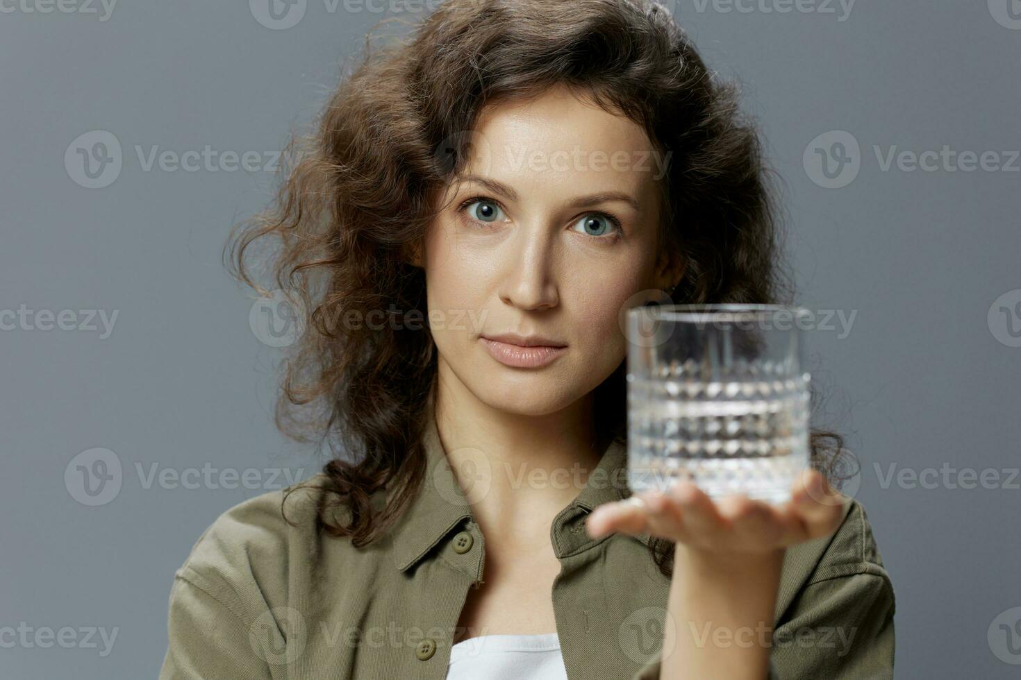 grave adorable Rizado hermosa mujer en casual caqui verde camisa sostiene sugiere vaso de agua posando aislado en terminado gris azul antecedentes. sano estilo de vida. agua es vida concepto. Copiar espacio foto