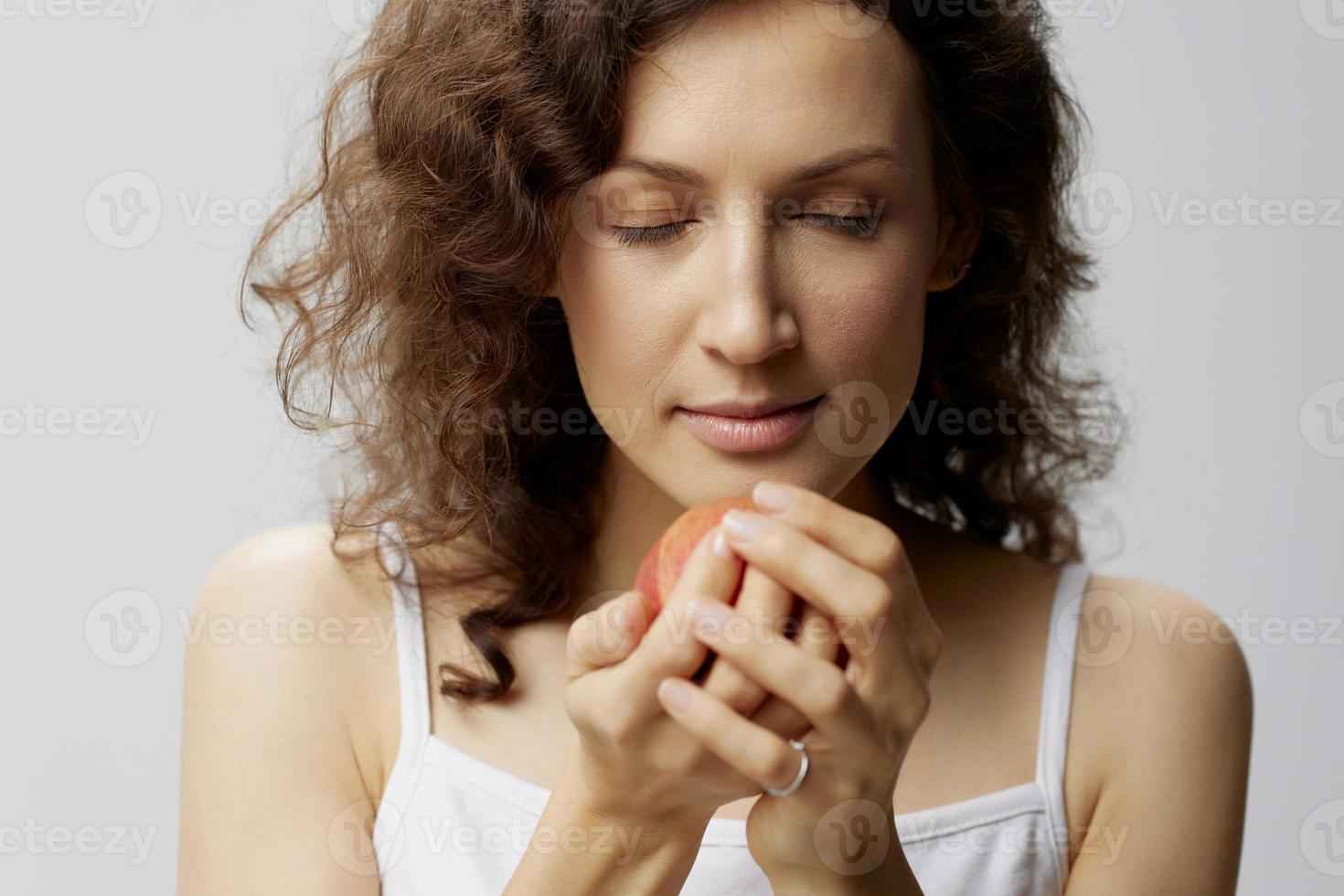 contento Rizado hermosa mujer en básico blanco camiseta olfateando un manzana disfruté oler de Fruta posando aislado en terminado blanco antecedentes. natural Respetuoso del medio ambiente productos concepto. Copiar espacio foto