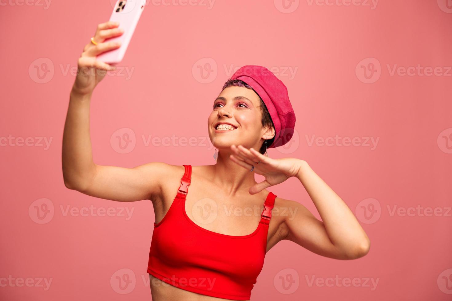 A young woman blogger with colored pink hair and a short haircut takes a picture of herself on the phone and broadcasts a smile in stylish clothes and a hat on a pink background monochrome style photo