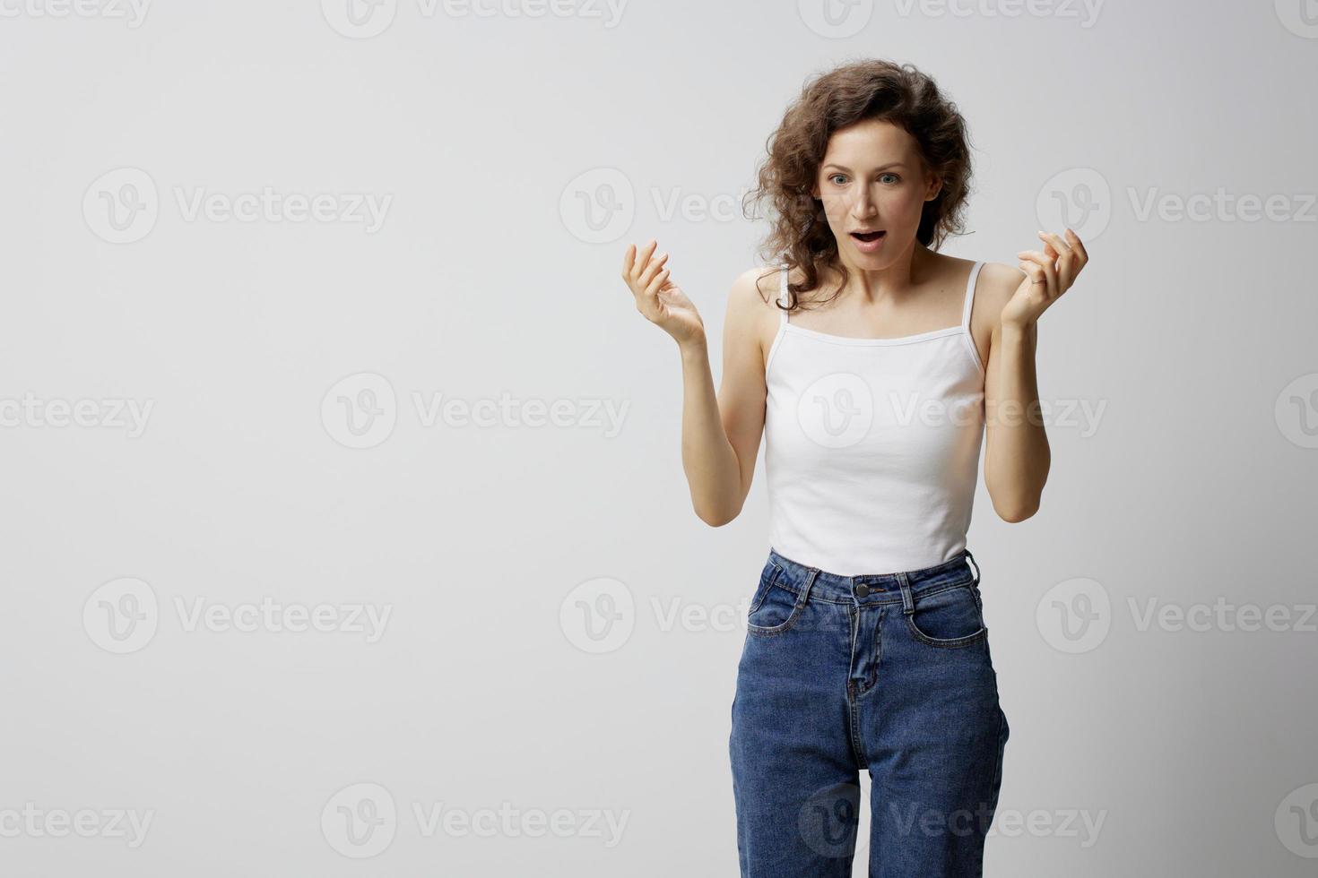 Confused shocked curly beautiful woman in basic white t-shirt raise hands up open mouth posing isolated on over white background. People Emotions Lifestyle. I can not believe it concept. Copy space photo