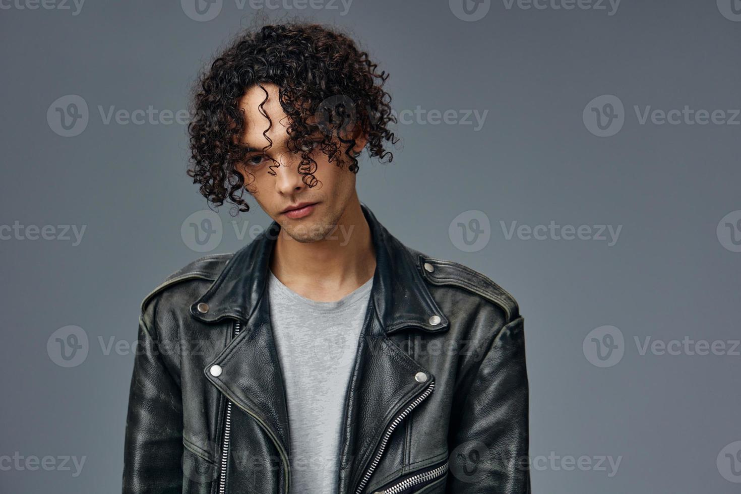 Self-confident stylish tanned curly man leather jacket looks at camera posing isolated on over gray studio background. Cool fashion offer. Huge Seasonal Sale New Collection concept. Copy space for ad photo