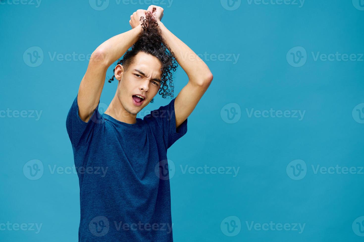 Positive joyful curly tanned Caucasian guy in basic t-shirt touches plays with hair curls posing isolated on over blue background. Lifestyle and Emotions concept. Good offer with copy space for ad photo