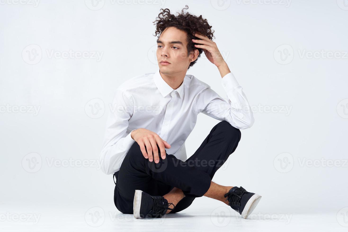 a guy in trousers in sneakers and a shirt sits on the floor and gestures with his hands in full growth photo