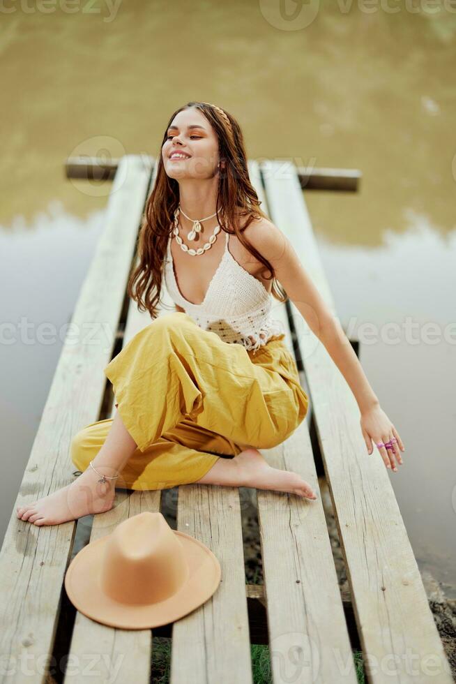 un hippie mujer se sienta con su sombrero en un puente por un lago con su manos arriba en el aire en un naturaleza viaje y sonriente felizmente en ropa ecológica. relajado estilo de vida foto