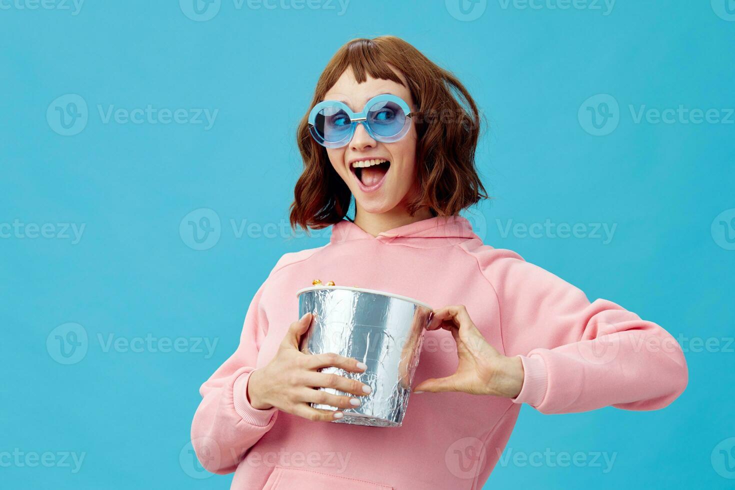 película concepto. contento alegre linda pelirrojo dama en rosado capucha Gafas de sol con palomitas de maiz posando aislado en azul estudio antecedentes. Copiar espacio bandera. Moda cine. entretenimiento oferta foto