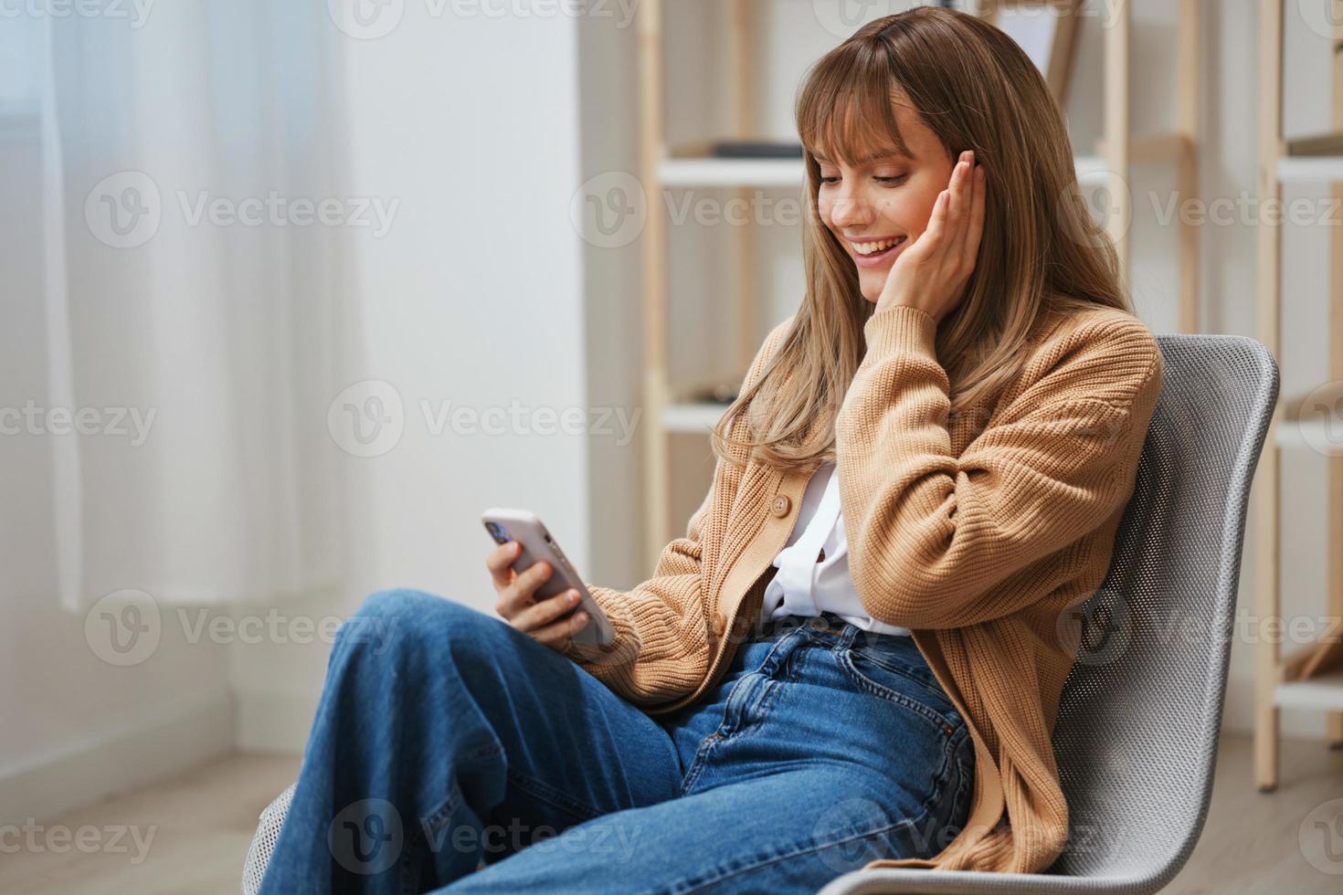 Cheerful young blonde woman in warm sweater get good news on studies by phone touch cheek sitting in armchair at home. Pause from work, take a break, social media in free time concept. Copy space photo