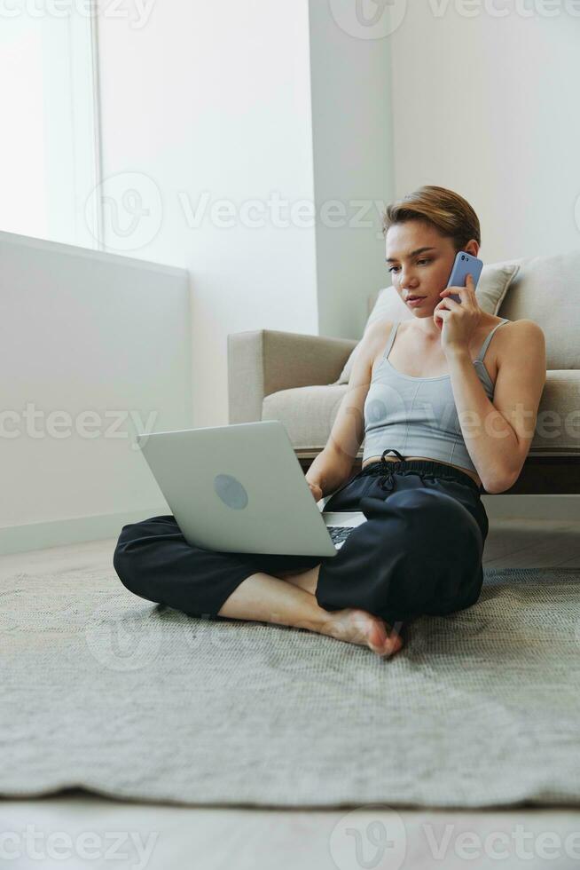 Freelance woman with laptop and phone works from home sitting on the floor in her home clothes with a short haircut, free copy space photo