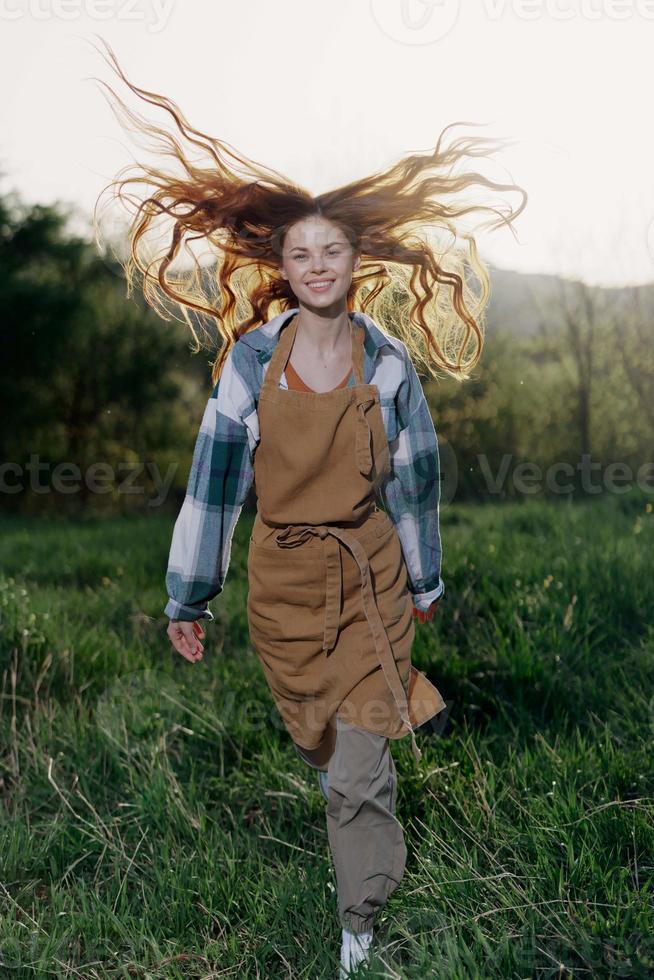 mujer caminando en el césped en naturaleza sonriente en granja ropa con hermosa pelo volador en el viento contento en el rayos de verano puesta de sol ligero foto