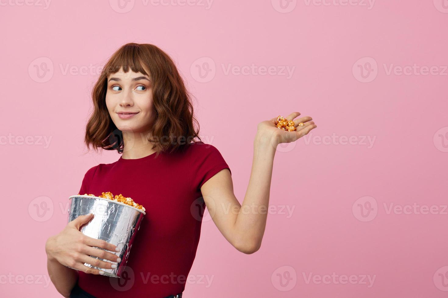 emocionado confuso linda pelirrojo dama en rojo camiseta con palomitas de maiz Listo para película noche posando aislado en terminado rosado estudio antecedentes. Copiar espacio bandera. Moda cine concepto. entretenimiento oferta foto