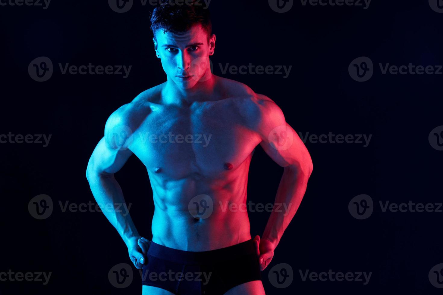 sexy hombre atleta en pantalones cortos sostiene manos en su cinturón en un negro antecedentes foto