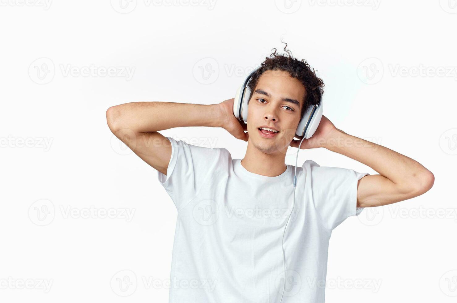 happy curly-haired guy in headphones and in a white t-shirt listens to music and holds his hands near his face photo