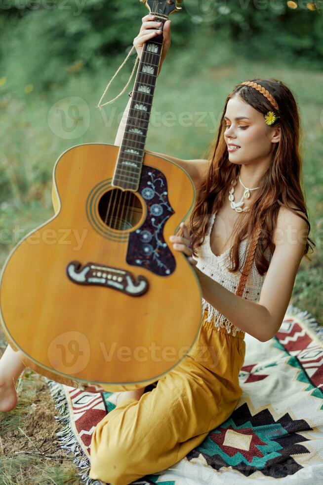 Hippie woman eco friendly playing guitar smiles and singing songs in nature sitting on a plaid by the lake in the evening in the sunset sunlight. A lifestyle in harmony with the body and nature photo