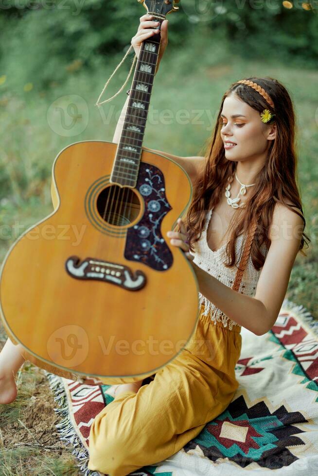 hippie mujer eco simpático jugando guitarra sonrisas y canto canciones en naturaleza sentado en un tartán por el lago en el noche en el puesta de sol luz de sol. un estilo de vida en armonía con el cuerpo y naturaleza foto