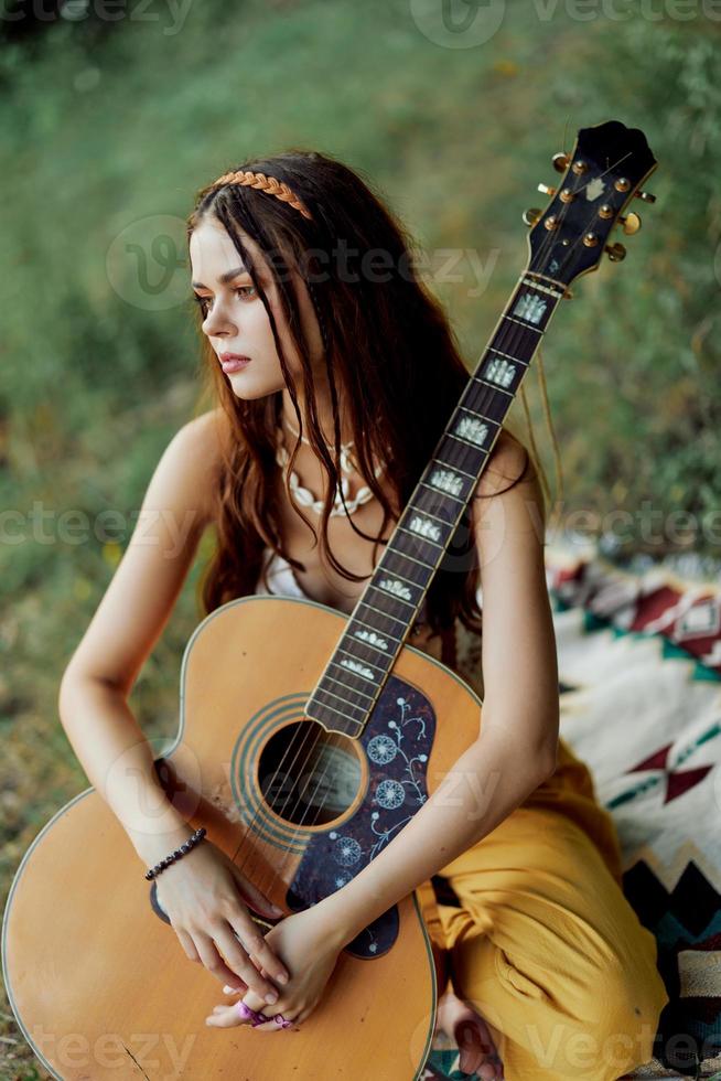 un niña en ropa ecológica hippie sentado con un guitarra y mirando a un puesta de sol en el verano foto