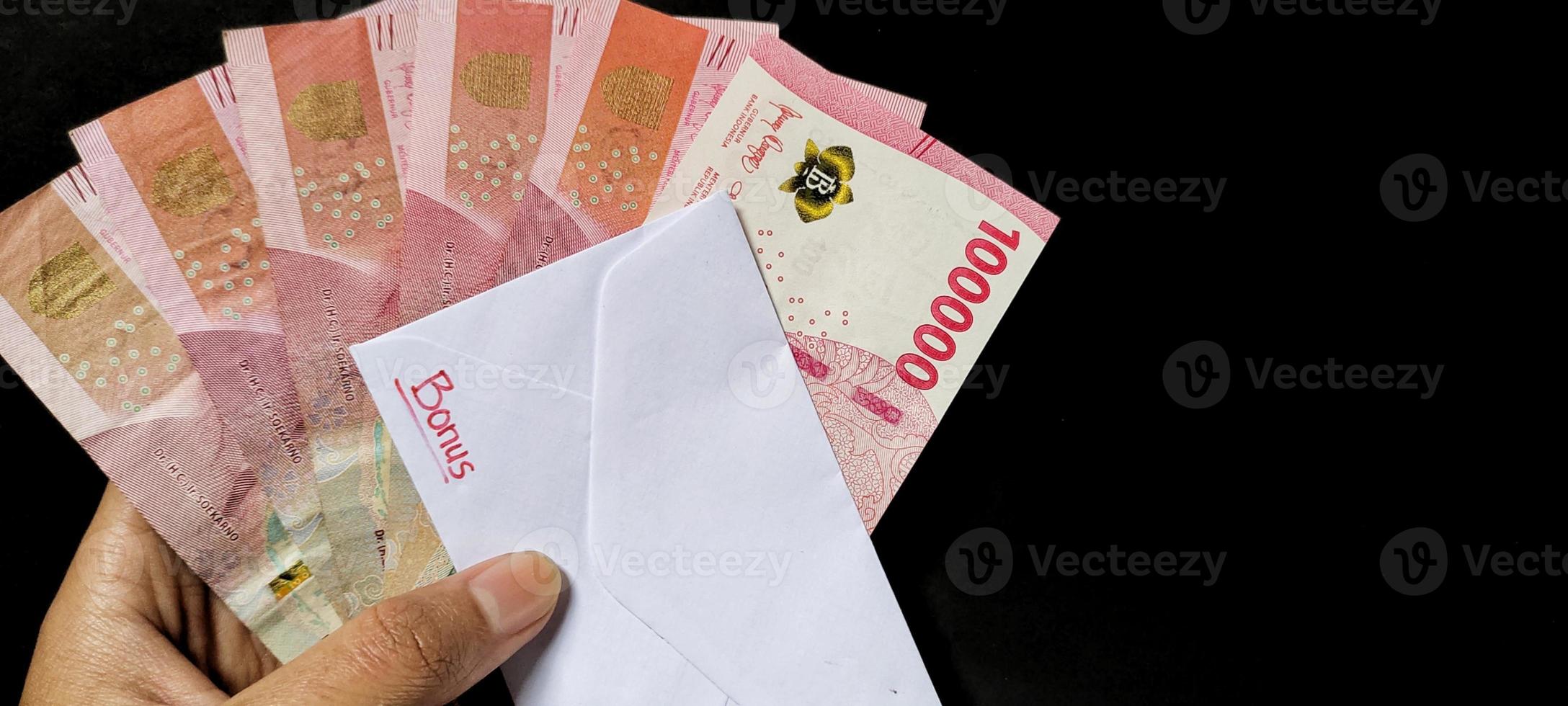 Man holding a white envelope written of Bonus and new Indonesian banknotes, usually Tunjangan Hari Raya or called THR are given to employees ahead of Eid. Isolated on black background photo