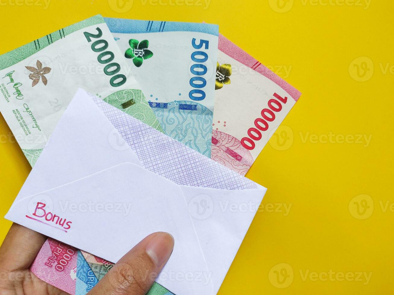 Man holding a white envelope written of Bonus and new Indonesian banknotes, usually Tunjangan Hari Raya or called THR are given to employees ahead of Eid. Top view photo