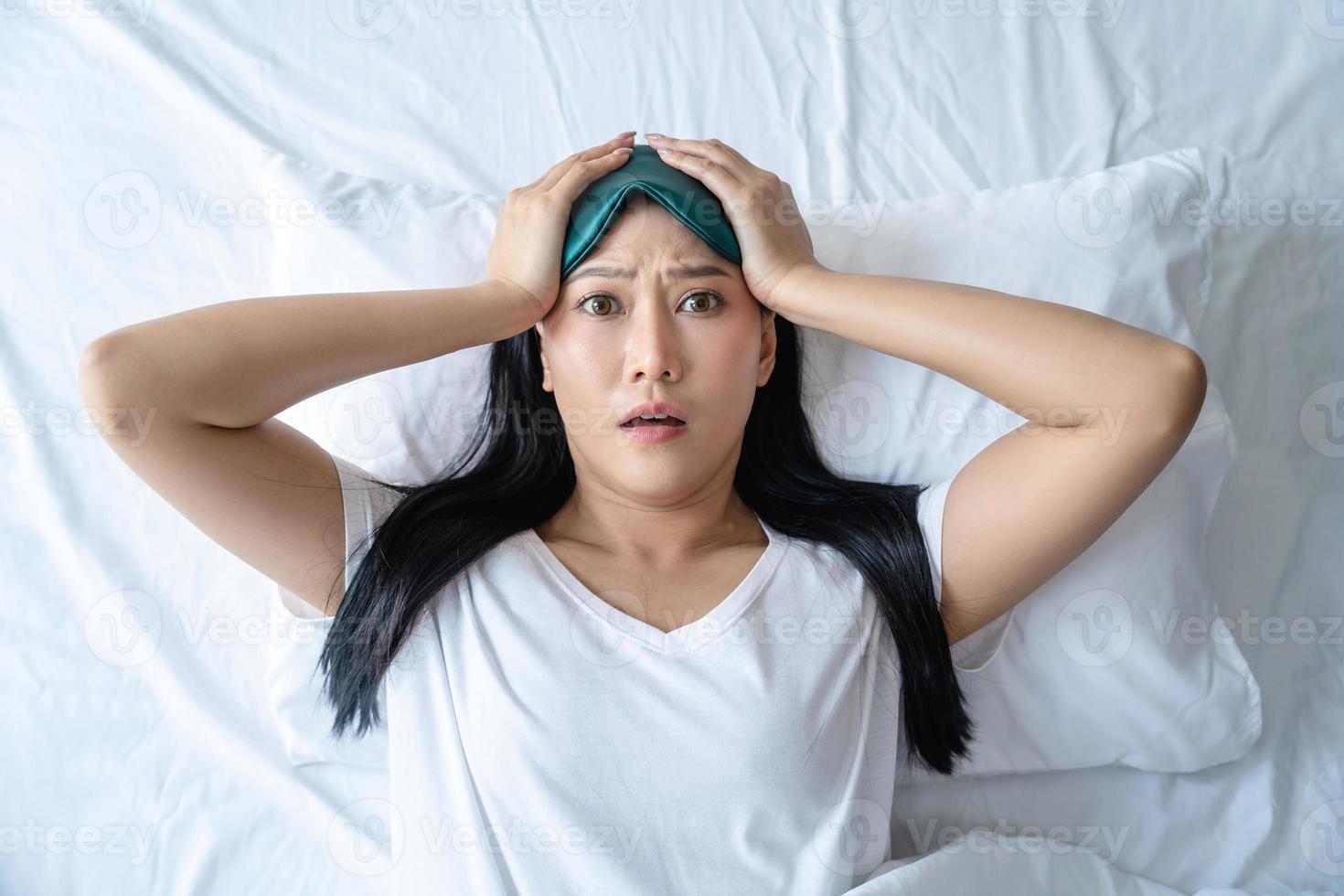 Close-up shot of panicking asian cute girl lying in bed on pillow in sleeping mask and pajamas. She had a bad dream. photo