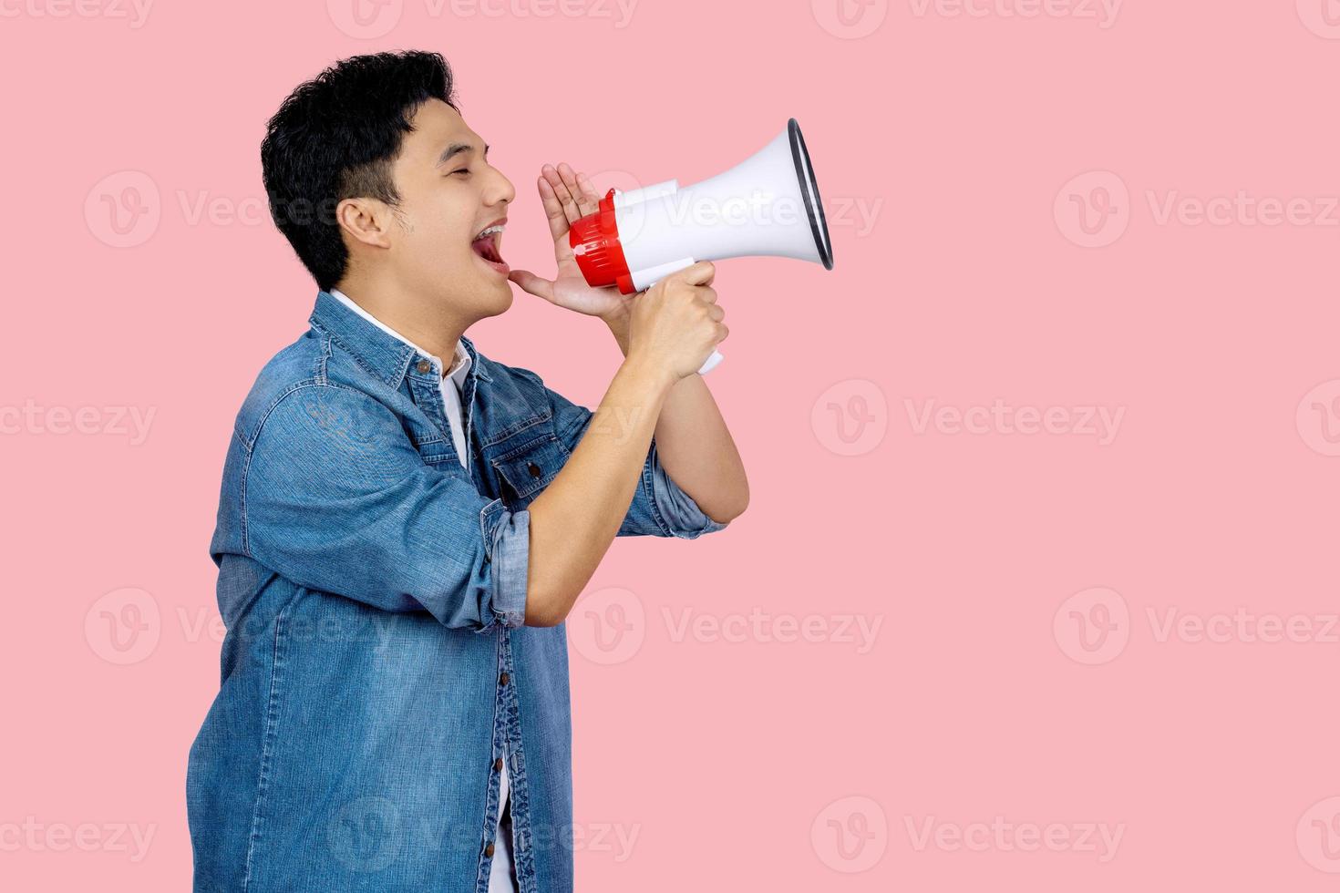 Happy young Asian man in blue shirt shouting announce into megaphone isolated on pink background in studio. photo