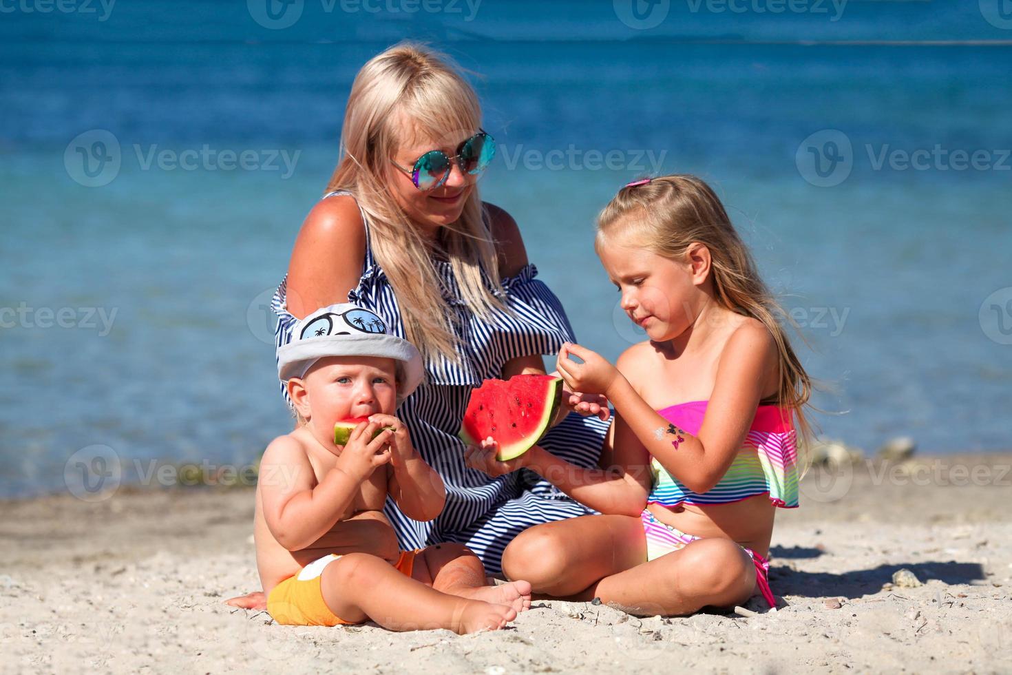 mamá y niños son sentado en el playa cerca el mar y comiendo sandía. vacaciones. foto