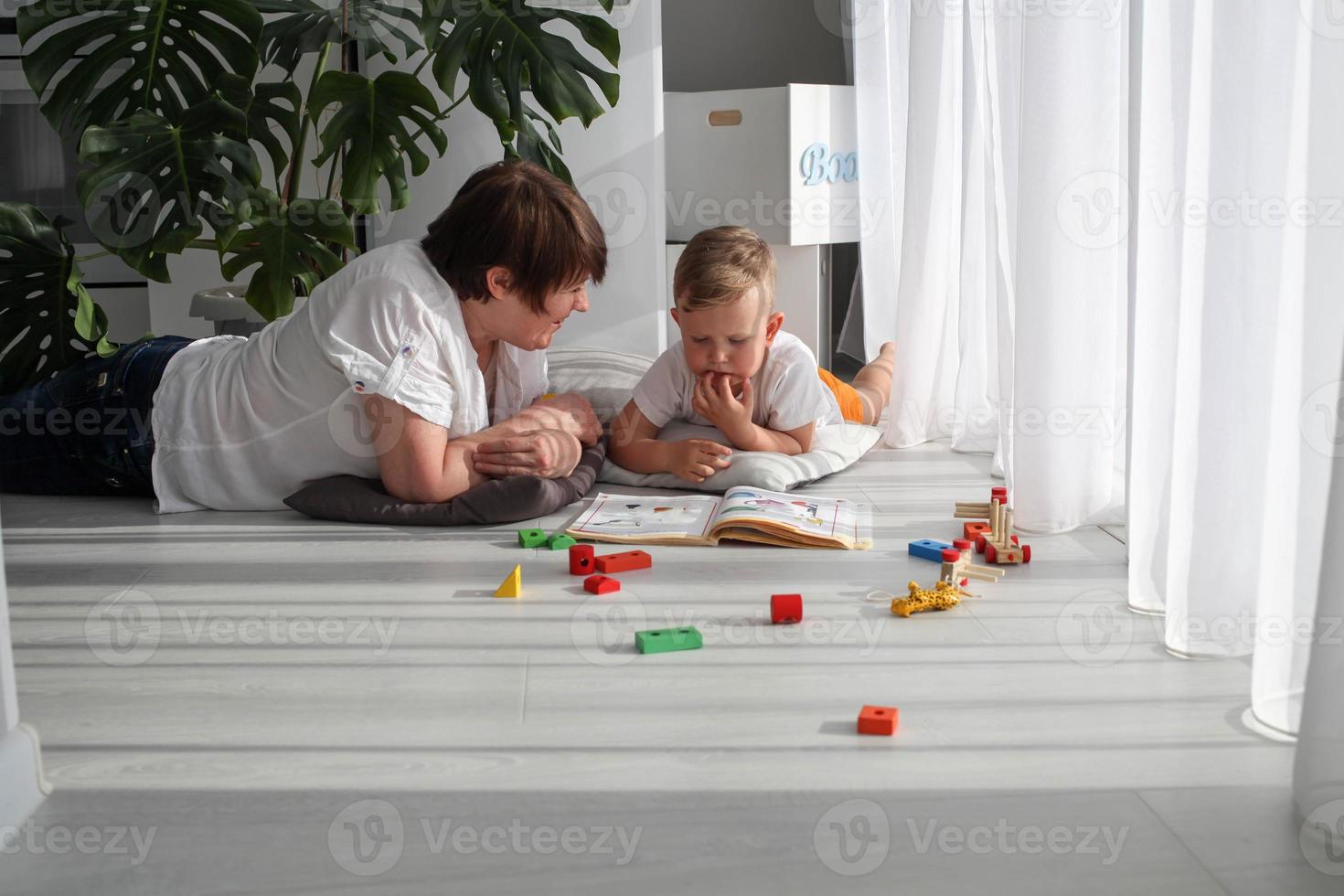 Grandmother is lying on the floor with her grandson and watching a book. The nanny is engaged in developing tasks with a toddler boy. Early development. photo
