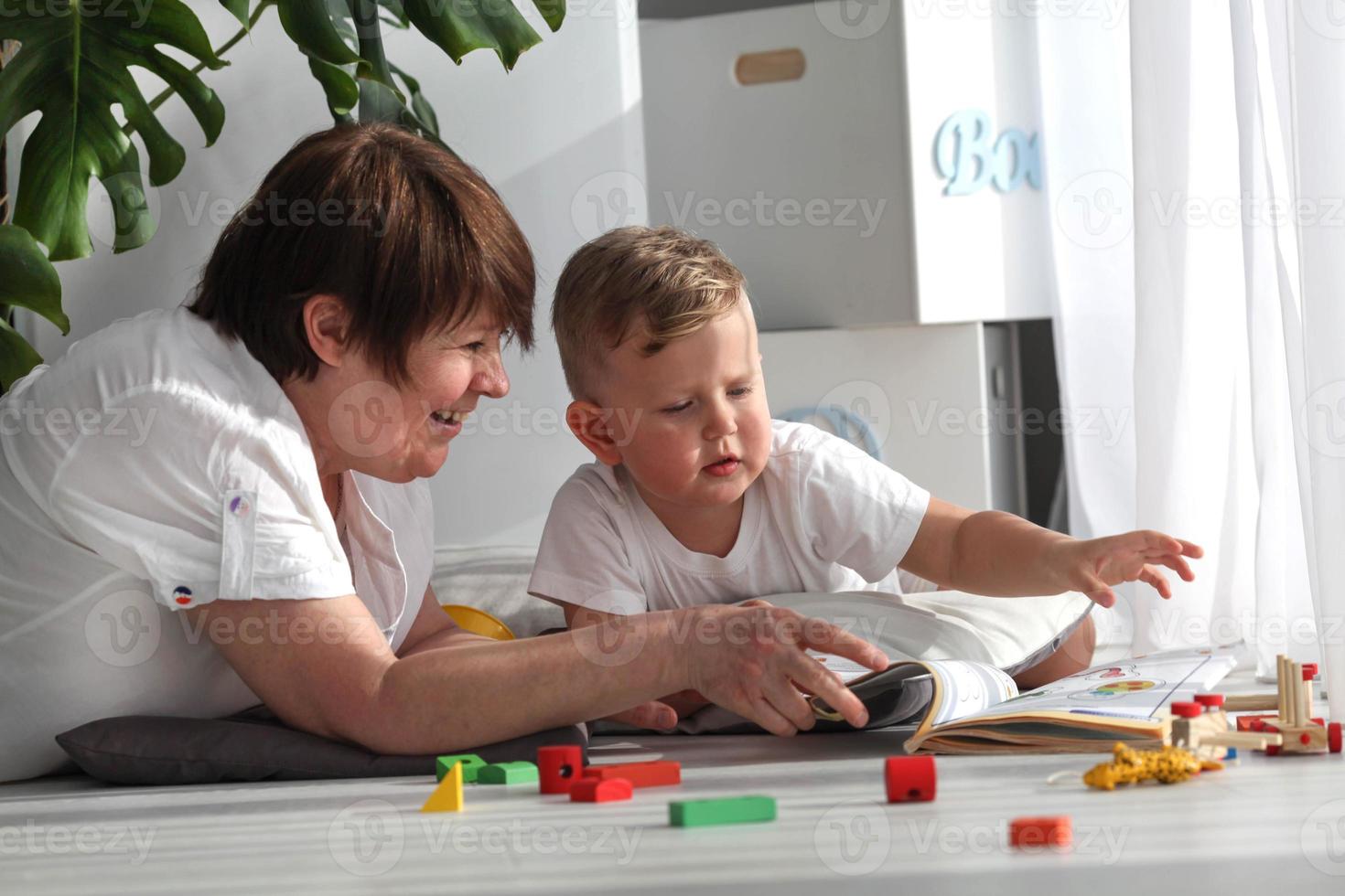 Grandmother is lying on the floor with her grandson and watching a book. The nanny is engaged in developing tasks with a toddler boy. Early development. photo