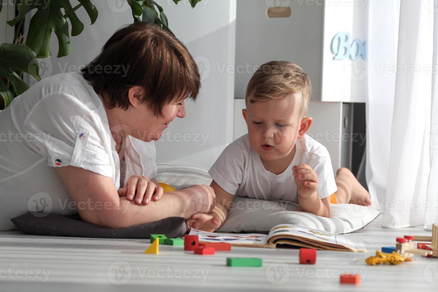 Grandmother is lying on the floor with her grandson and watching a book. The nanny is engaged in developing tasks with a toddler boy. Early development. photo