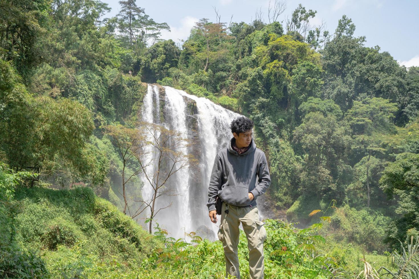 Man stand wear hoody in front of the great water fall with hand reel, Semarang central Java. The photo is suitable to use for adventure content media, nature poster and forest background.
