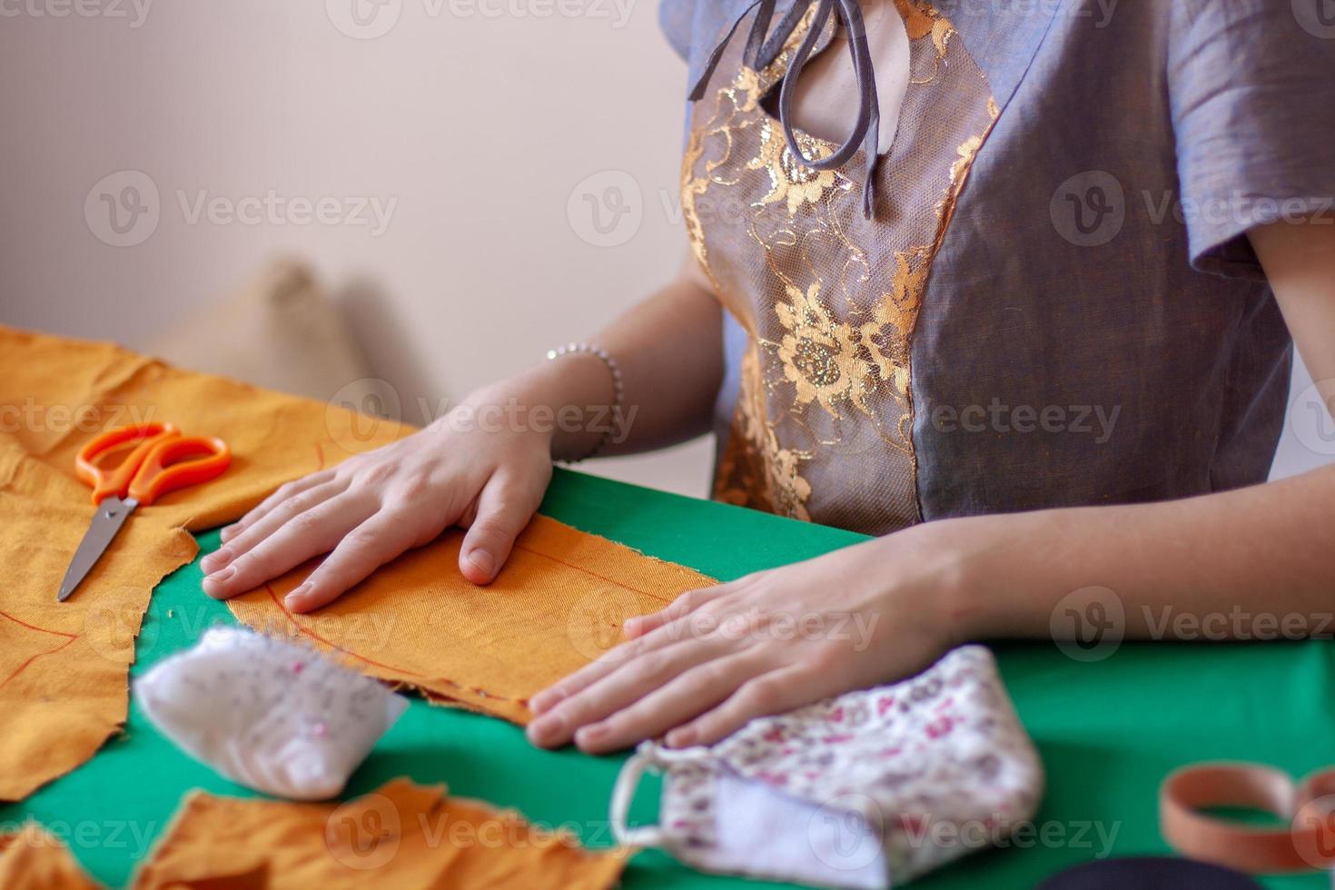 The seamstress smoothes the fabric blank for the mask. Fabric orange on a green table. Nearby are scissors and a finished mask. Focus on hands and tissue. Horizontal. photo