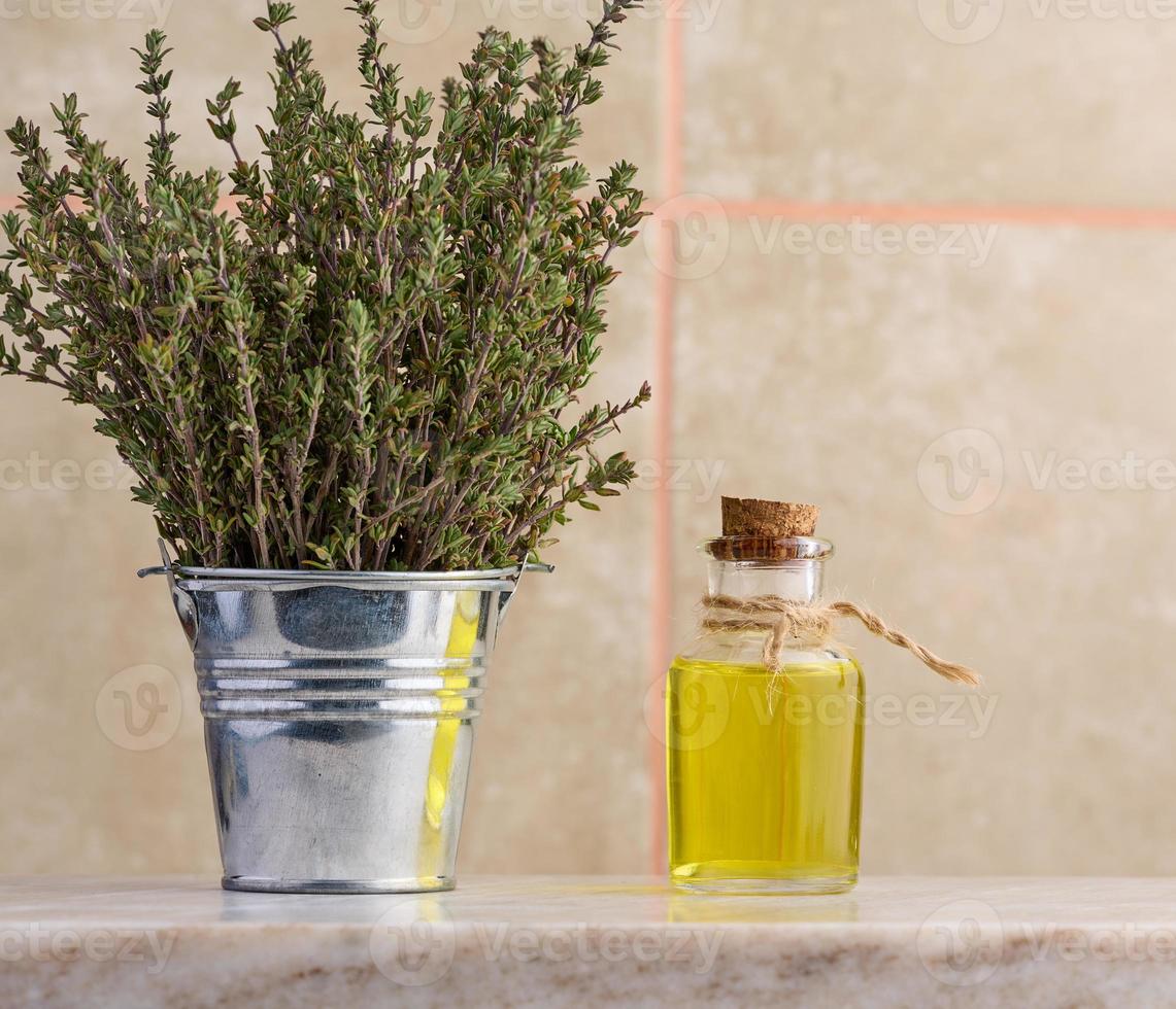 A clear glass bottle with oil and fresh thyme branches in a miniature bucket on a wooden table, cosmetic procedure oil photo