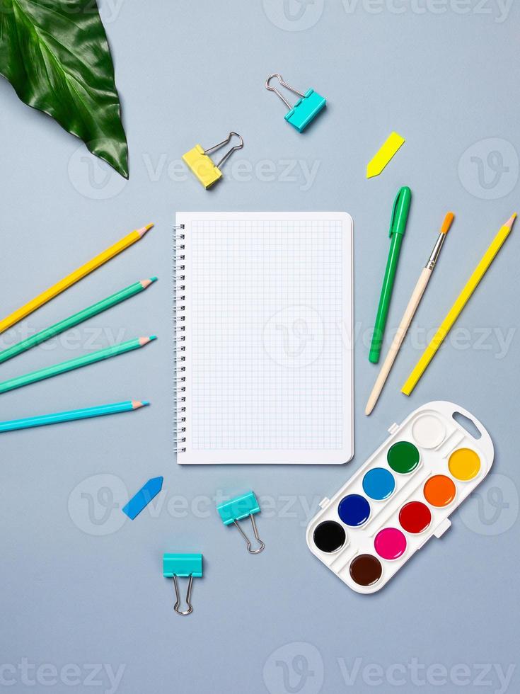 Top view arrangement with notepad with white sheets and pens, pencils, paper clips and other office supplies on blue background photo