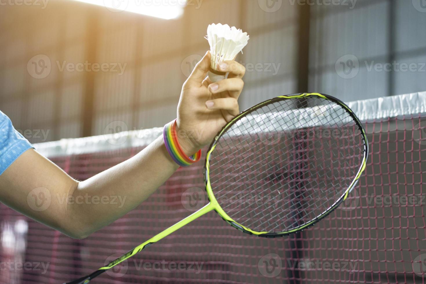 Badminton player wears rainbow wristbands and holding racket and white shuttlecock in front of the net before serving it to player in another side of the court, concept for LGBT people activities. photo