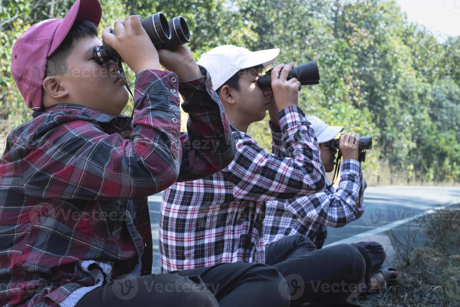 asiático Niños en tartán camisa usa gorra y participación un prismáticos, sentado en pavimento en local nacional parque a observar aves en árbol ramas y en cielo y a reloj insectos en árbol hojas. foto