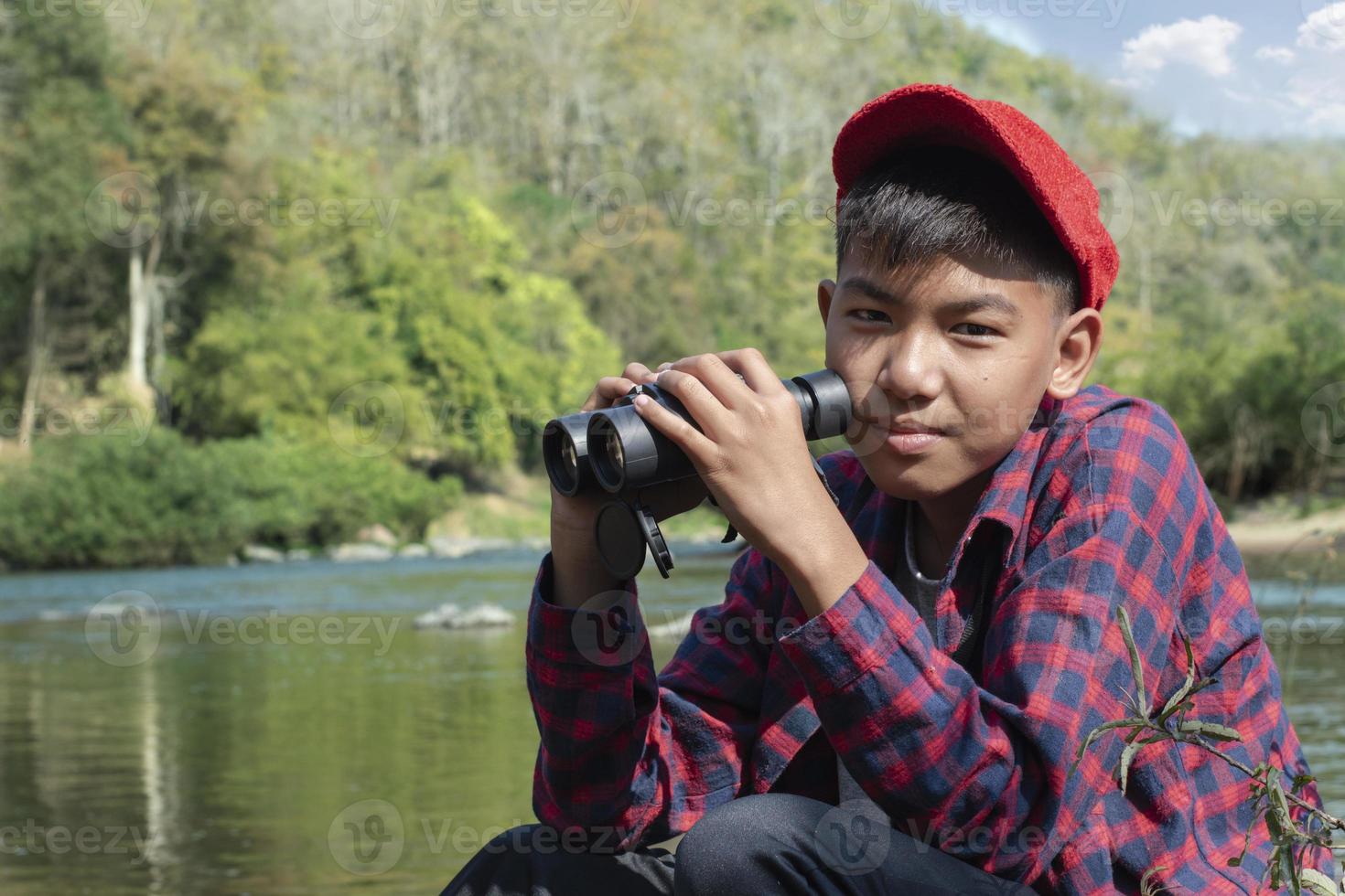 Asian boy in red cap and plaid shirt holds binoculars and sitting on rock in the middle of river in local national park to observe fish, nature and to watch birds on tree branches and flying on sky. photo