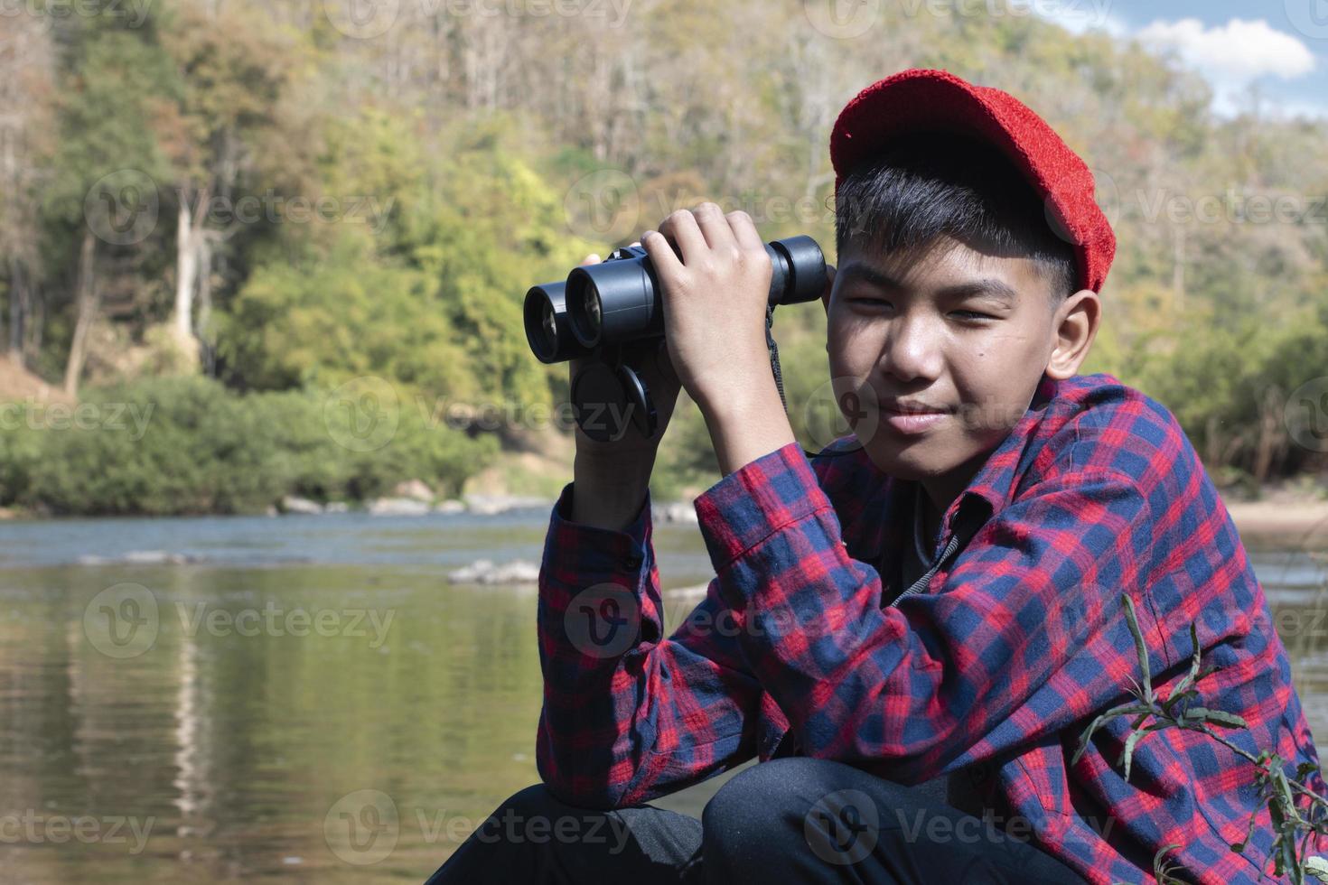 Asian boy in red cap and plaid shirt holds binoculars and sitting on rock in the middle of river in local national park to observe fish, nature and to watch birds on tree branches and flying on sky. photo