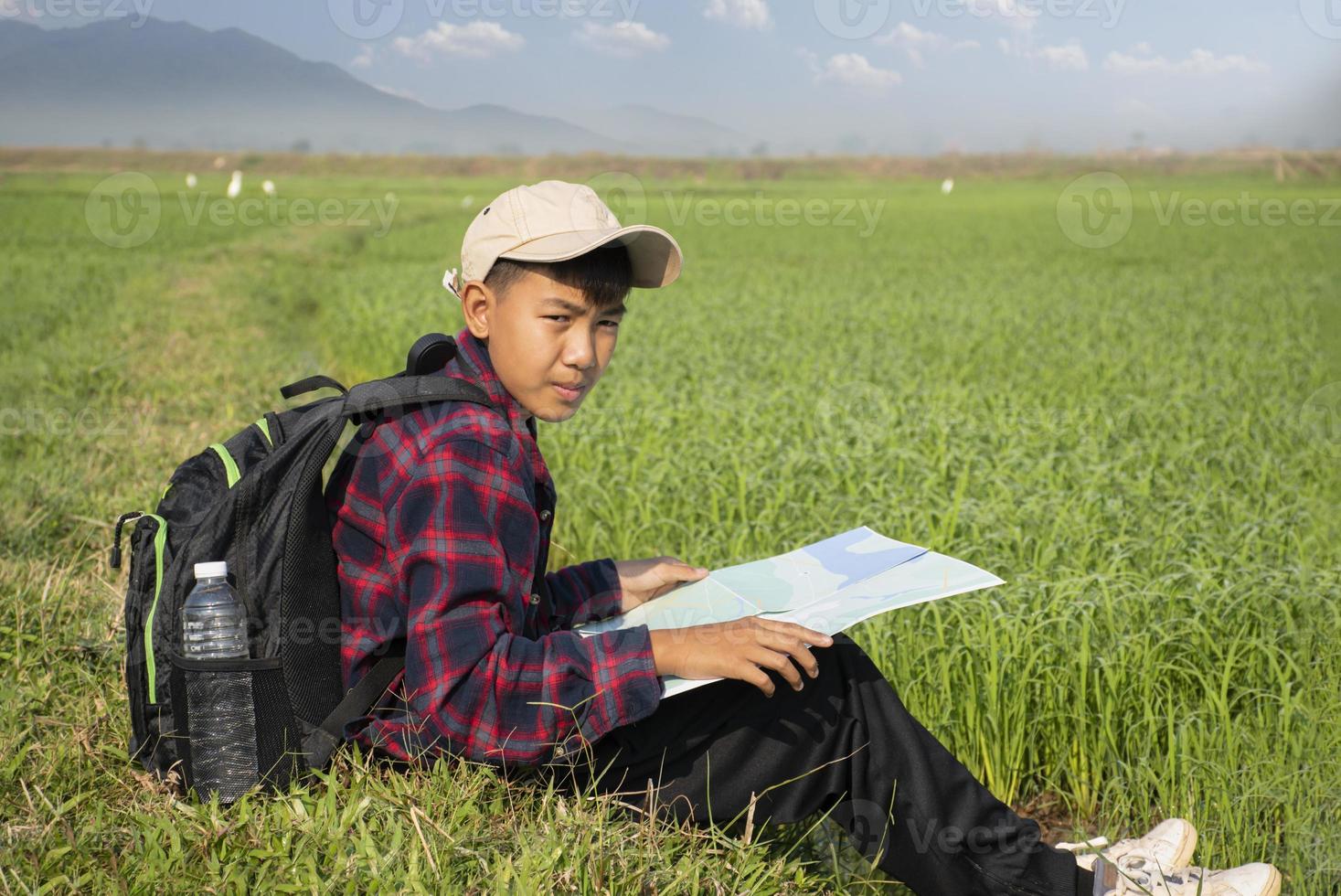 Asian boy wearing plaid shirt and a cap sitting on ridge of rice paddy field, holding a map and a binoculars, reading map before observing birds, pm 2.5 smoke and farmland borders, soft focus. photo