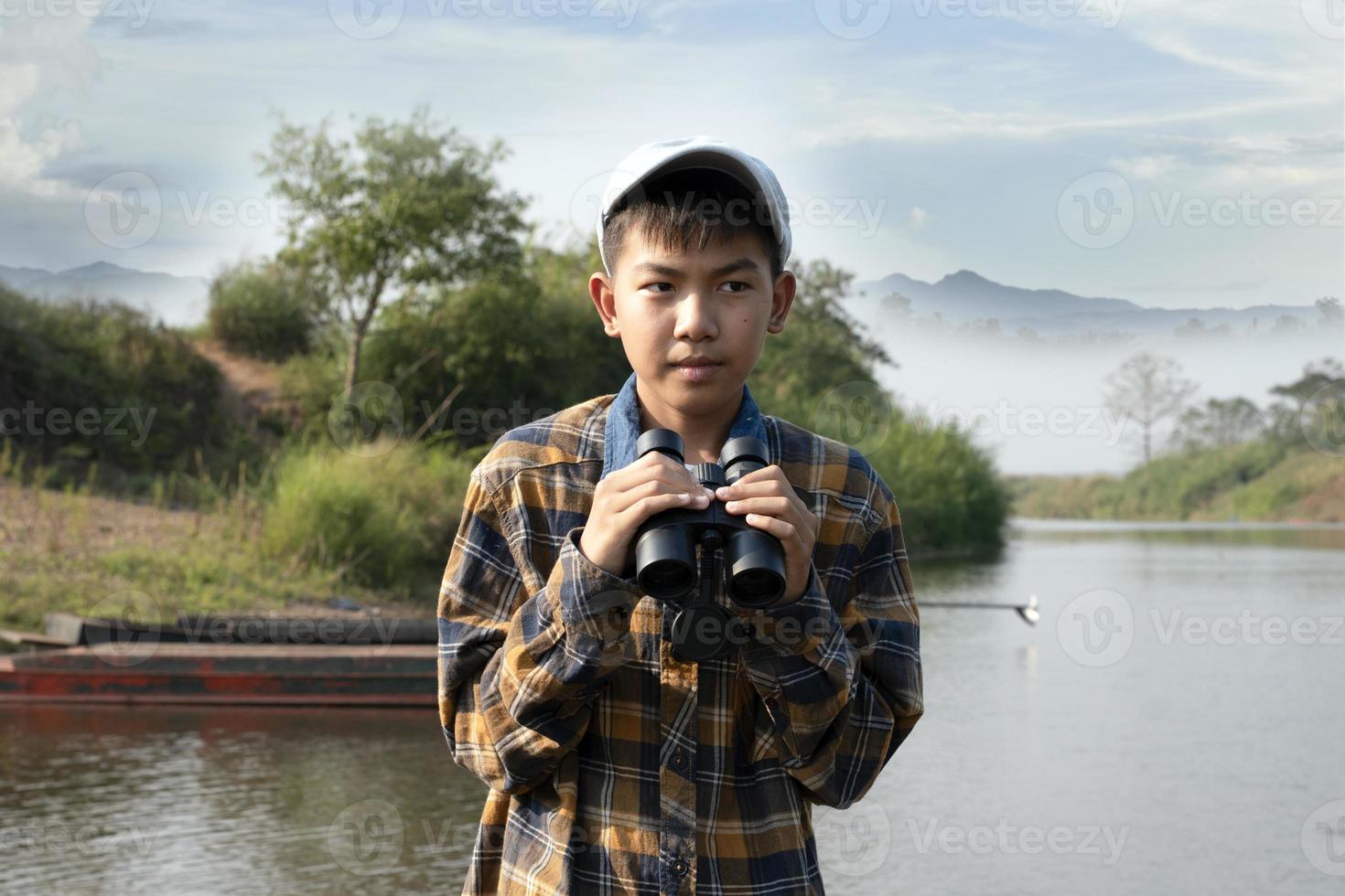 Asian boy in plaid shirt and cap, sitting and holding binoculars on bow of a boat parked beside a river to observe birds flying in the sky and fish swimming in the river during his summer holidays. photo