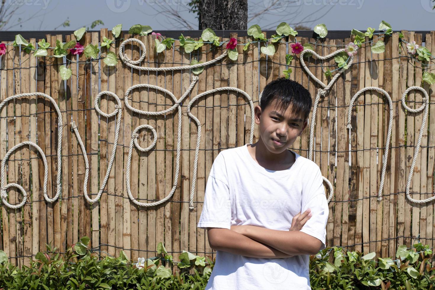 Cute asian boy in white t-shirt, standing with arms folded in front of a bamboo wall that has Thai characters on it, which means 'in love' in English. photo