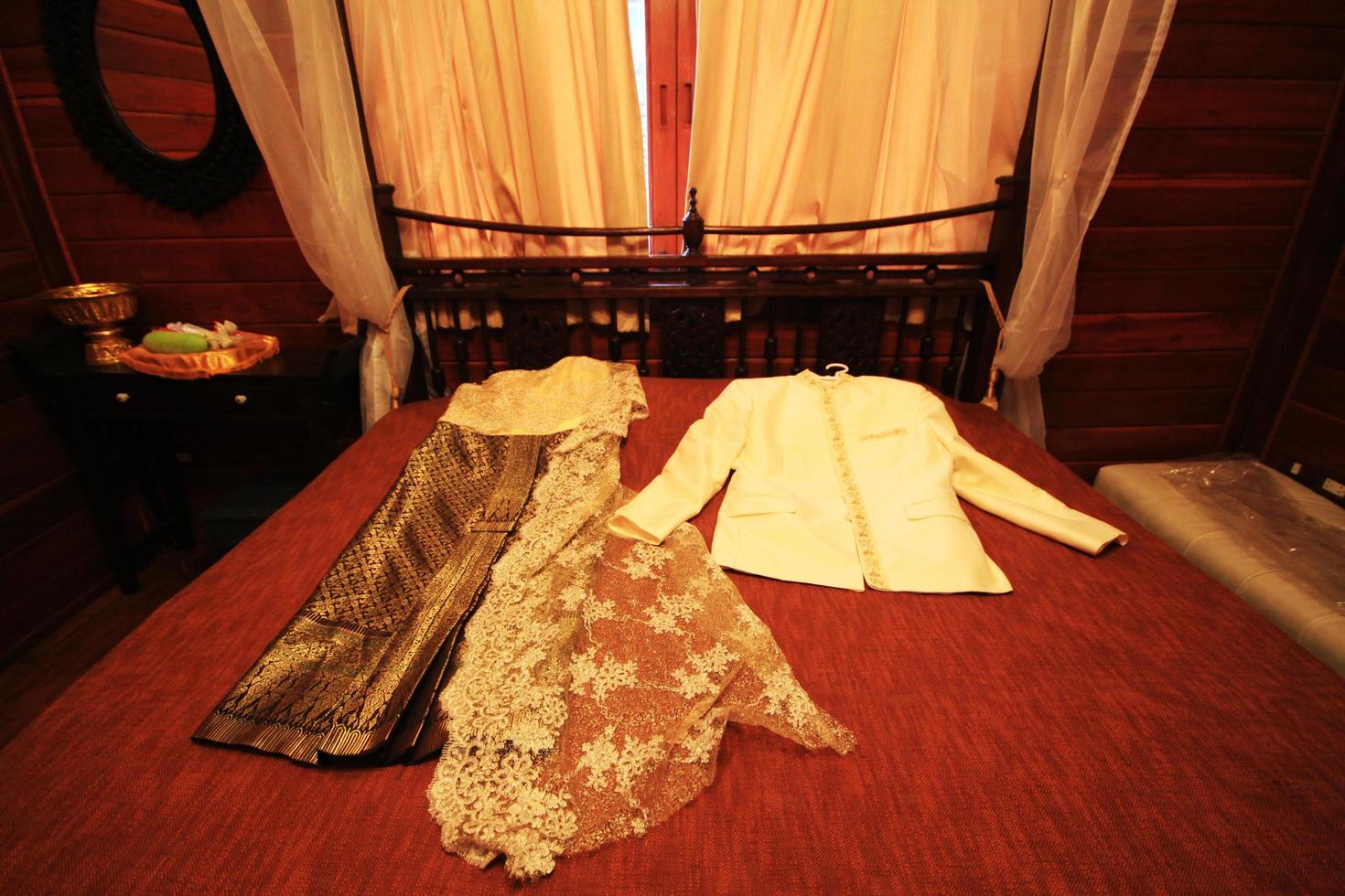 tailandés Boda vestir y traje acostado en cama en tailandés Boda ceremonia. novio y novia ropa concepto. foto
