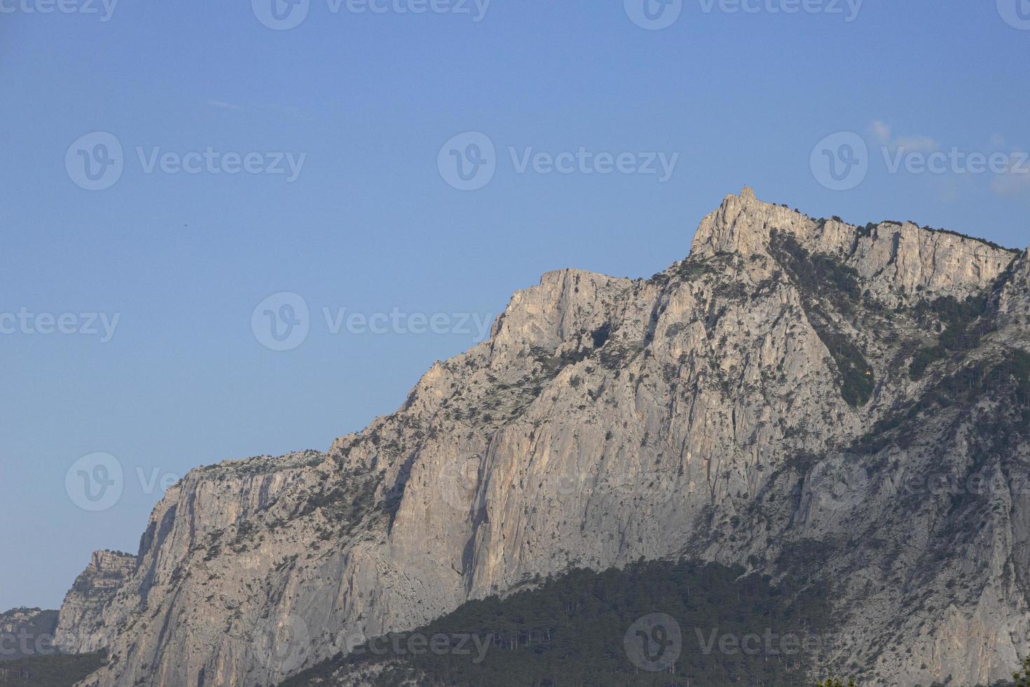 The peak, high gray cliffs against the blue sky, the forest at the foot of the mountains. Mountain scenery. photo